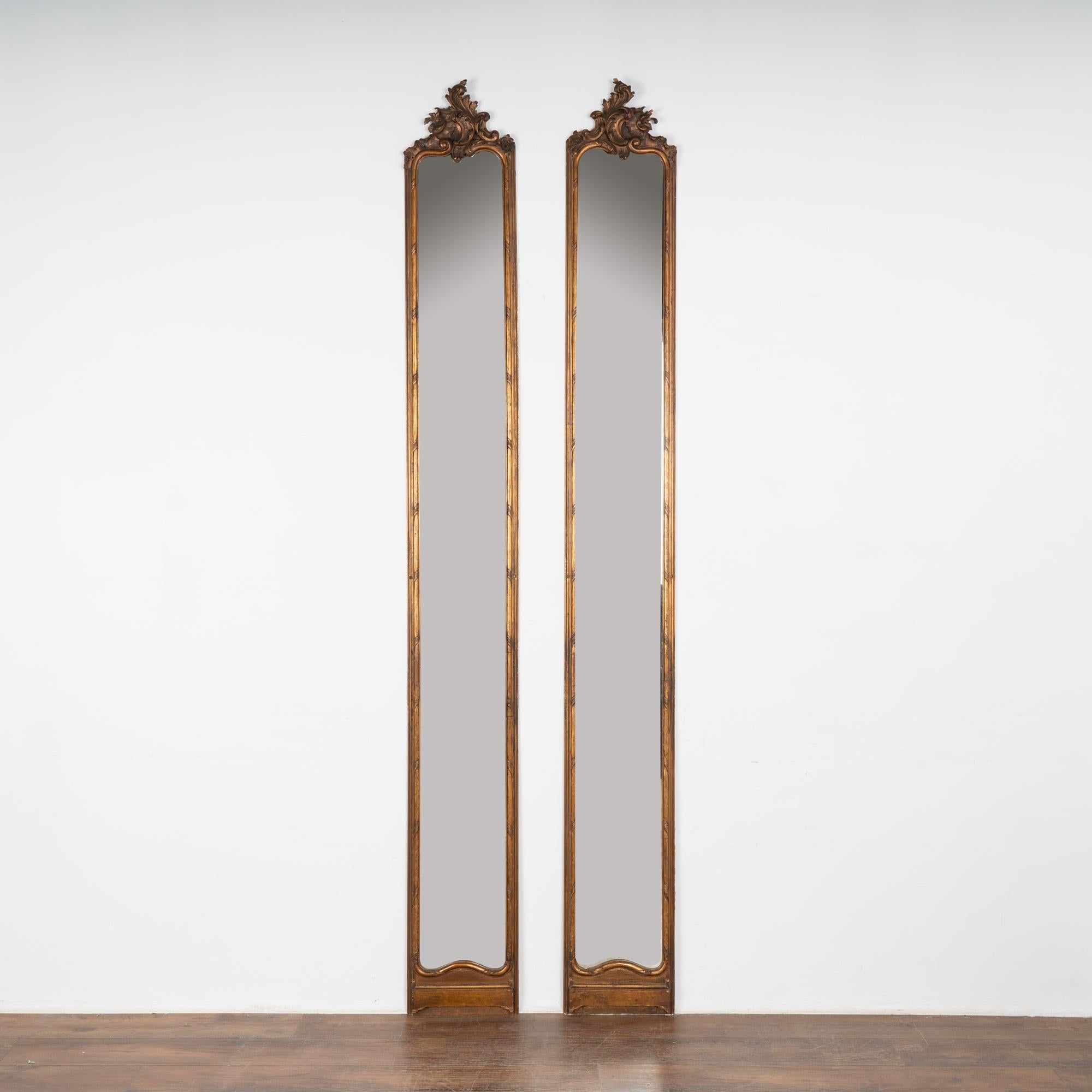 Rare pair of exceptionally tall, narrow mirrors crowned with carved flourish and bronze gilt finish.
Original distressed glass plates, still usable and elegant in length. Holes in each frame which prior owners used to hang each.
All scratches,