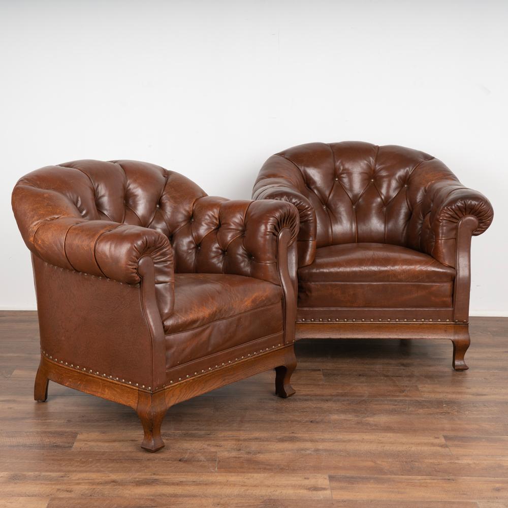 Pair, vintage leather barrel back club chairs with oak wood frame base and feet.
Good looking vintage brown leather, nailheads and buttons.
Natural distress including scratchs, marks, dings reflect generations of use. One chair missing 3