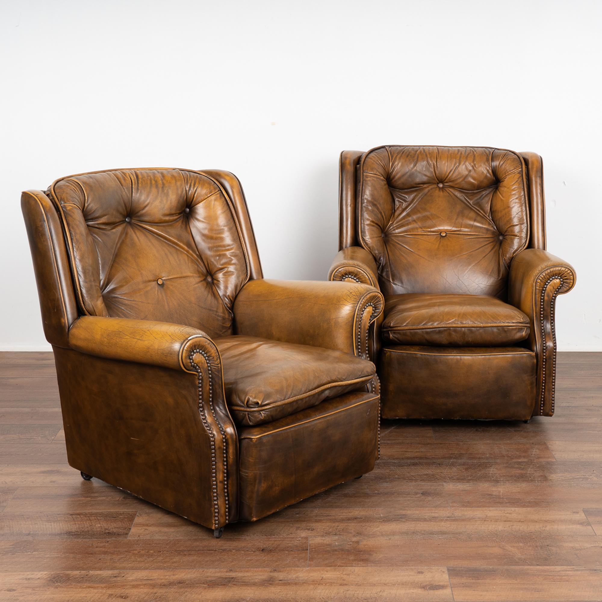 Pair, vintage leather lounge chairs with tufted back each resting on four castor feet.
Vintage brown leather with mottled dark and lighter tones, nail head trim along arms and six buttons along back cushion.
Natural distress including scratches,