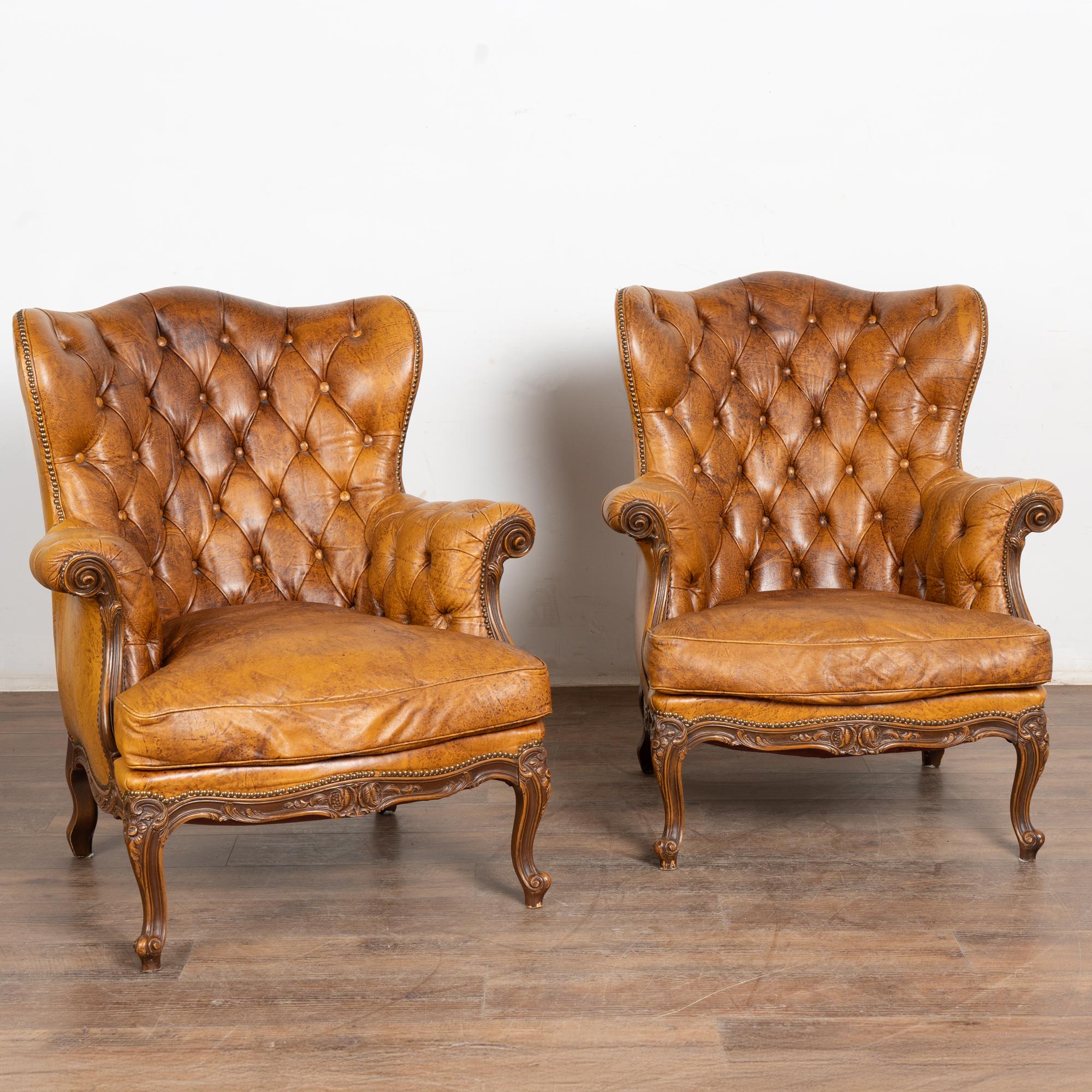 Pair, brown leather wingback club chairs in vintage brown leather. Decorative carved arms, skirt and cabriole legs.
Tufted back, cabriolet feet, buttons in place, at least one nail head trim missing. 
Vintage leather reflects typical age related
