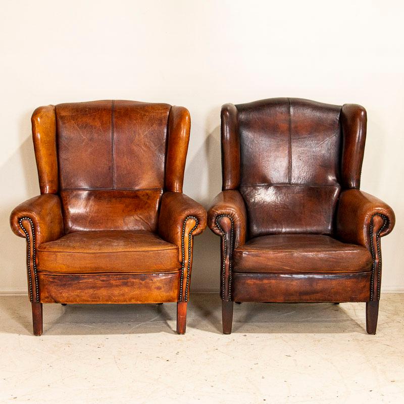 The warm, aged patina of the vintage leather invites one to sit and linger in this impressive pair of high backed club chairs. Notice that one has a darker patina than the other with nailheads and seams still intact on both. Both chairs show typical