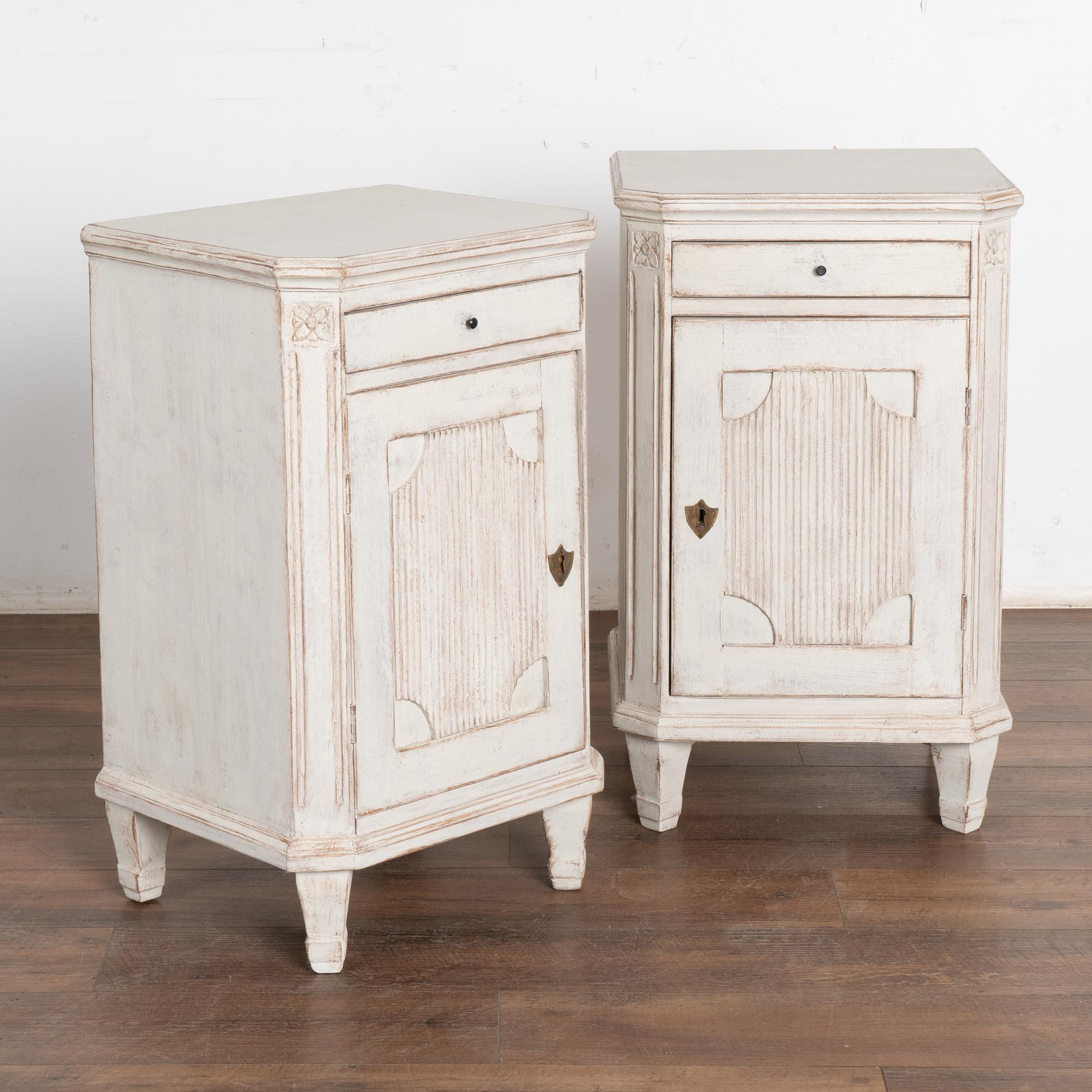 Pareja de mesitas de noche o pequeños armarios pintados de blanco, de estilo Gustaviano sueco, sobre patas cónicas.
Los lados inclinados tienen una sencilla talla acanalada rematada con un medallón floral. La talla estriada acentúa bien los paneles