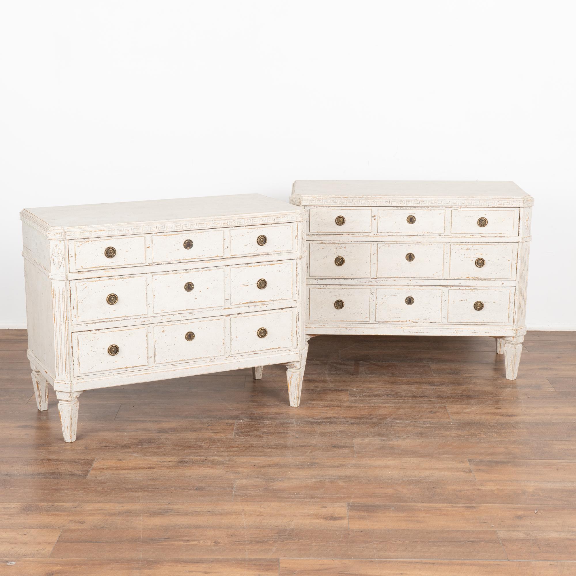 A pair of decorative Gustavian pine chest of drawers painted in shades of  white with Greek key motif carved in upper top molding.
Canted fluted side posts with upper carved floral medallion, raised panels along the three drawers, all setting on