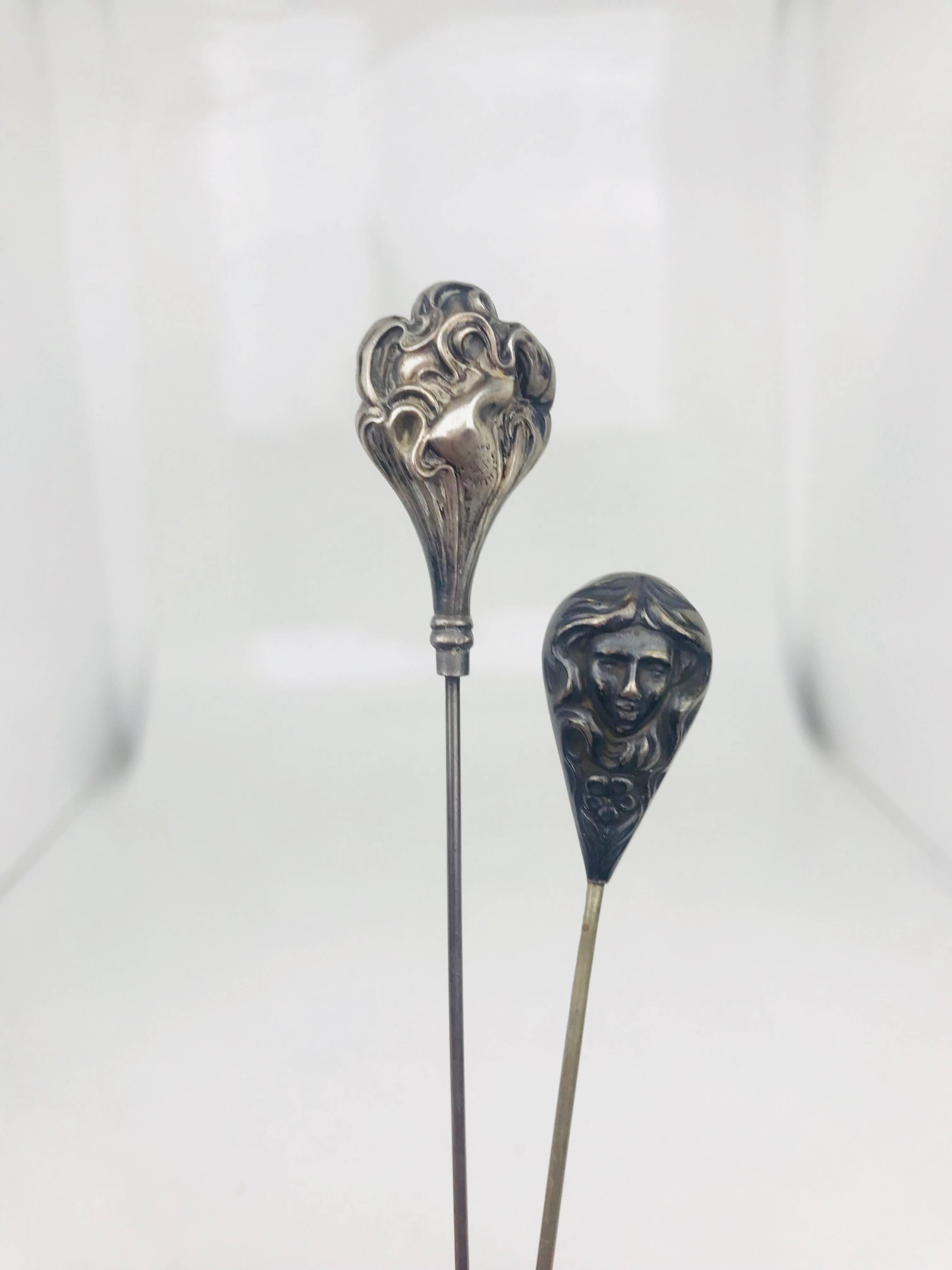 Victorian Sterling Silver Hat Pin Pair feature Pear-Shaped, Stylized, Hollow Heads.
Circa 1890
 
The smaller hat pin depicts a woman's head and face with long, flowing hair and a flower beneath the chin.  It is two-faced, identical on both sides.