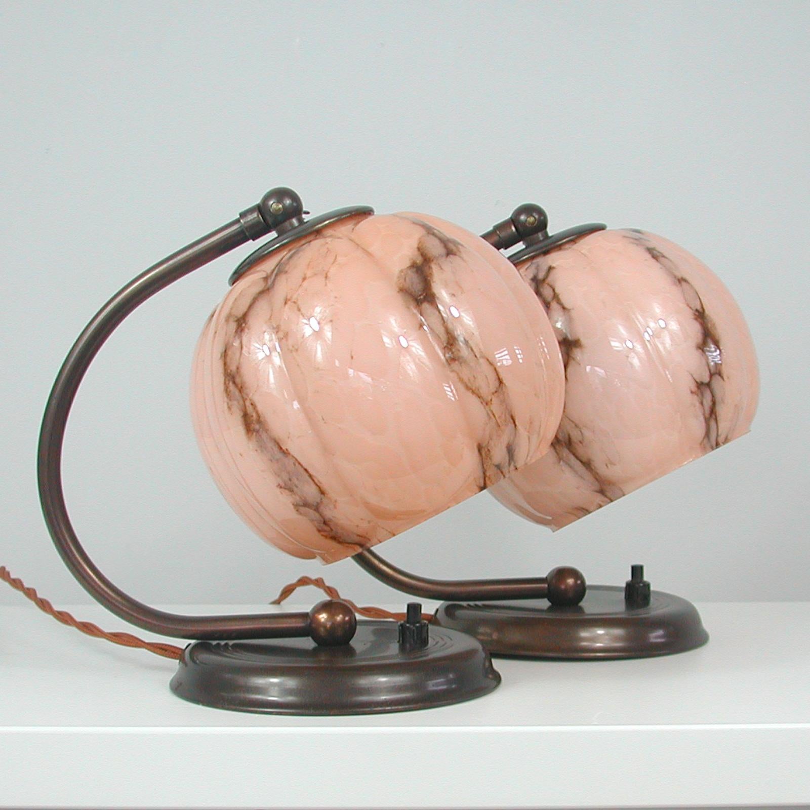 These unusual table or bedside lamps were designed and manufactured in Germany in the 1930s during the Art Deco / Bauhaus period. They are made of bronzed brass and have got pale pink / salmon colored marbled opaline glass lamp shades. The lamps