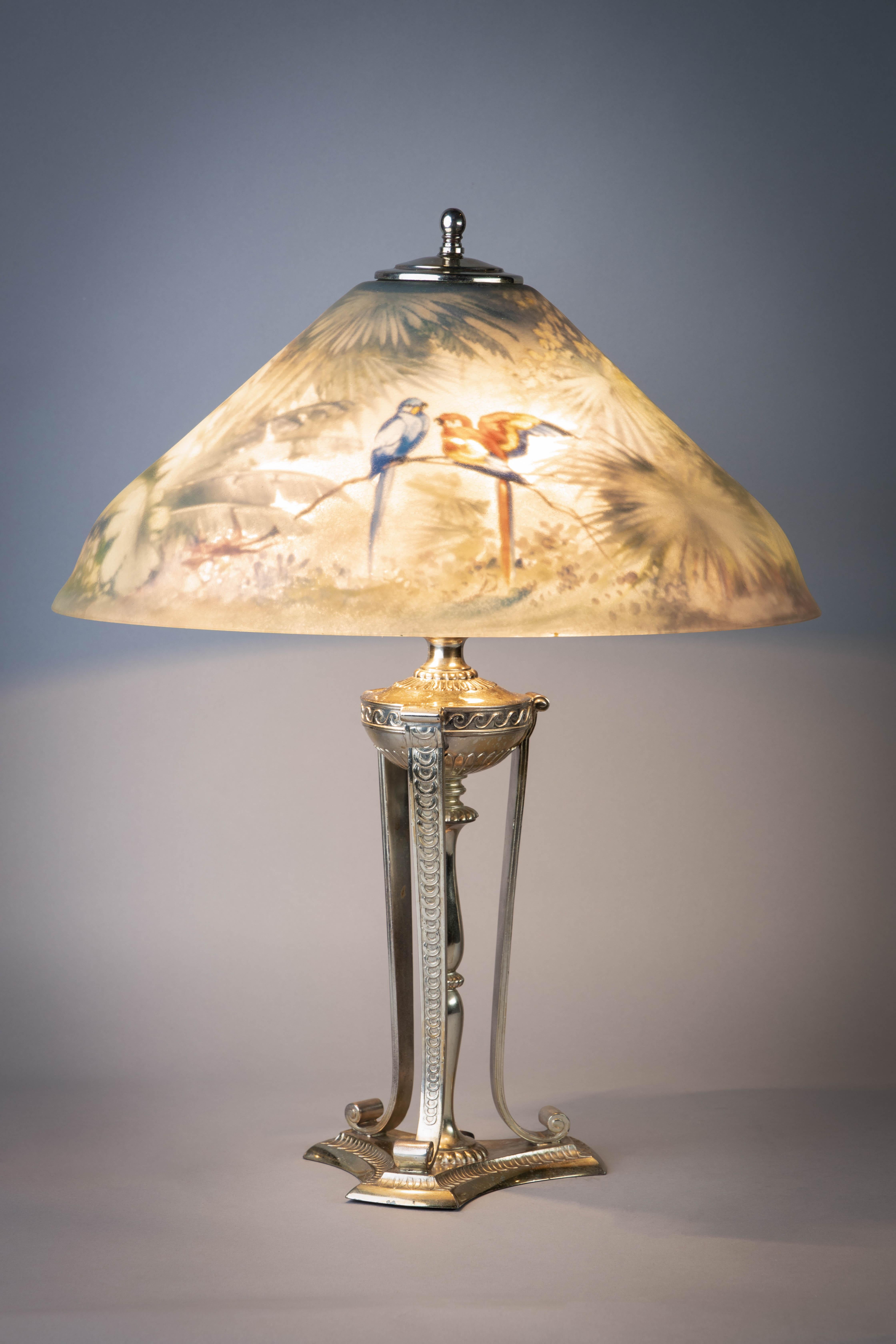 The conical shade decorated with birds perched on tropical branches, three-light socket, on a neoclassical style urn and columnar standard flanked by three pillar supports ending in scrolling feet and a stepped triangular base.