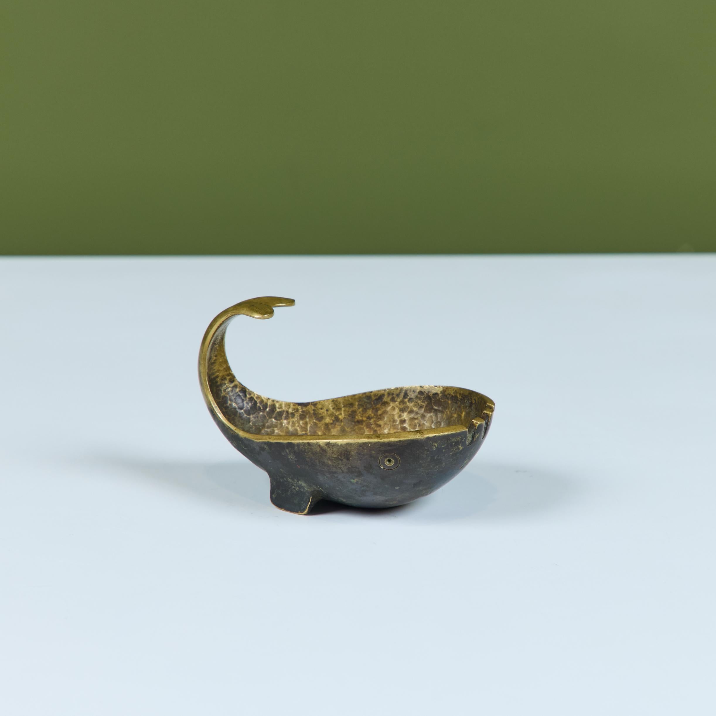 Cenicero de bronce en forma de ballena, con la cola curvada y tres ranuras en la parte delantera de la pieza. Tiene el interior martillado y una ligera pátina verdigris. Un sello en relieve en la parte inferior reza Pal-Bell Co. Fabricado en