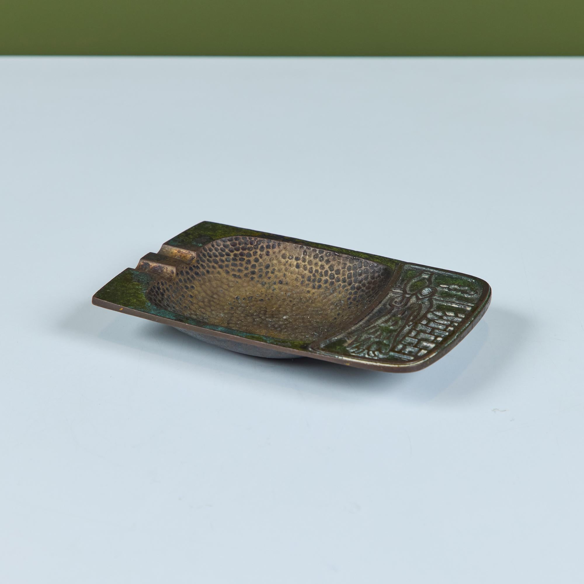 Mid-century brass Pal-Bell ashtray features a rectangular shape with a deep square hammered well and two cigarette notches. The opposite end showcases a figure and falcon. An embossed stamp on underside reads “Pal-Bell Co. Made in