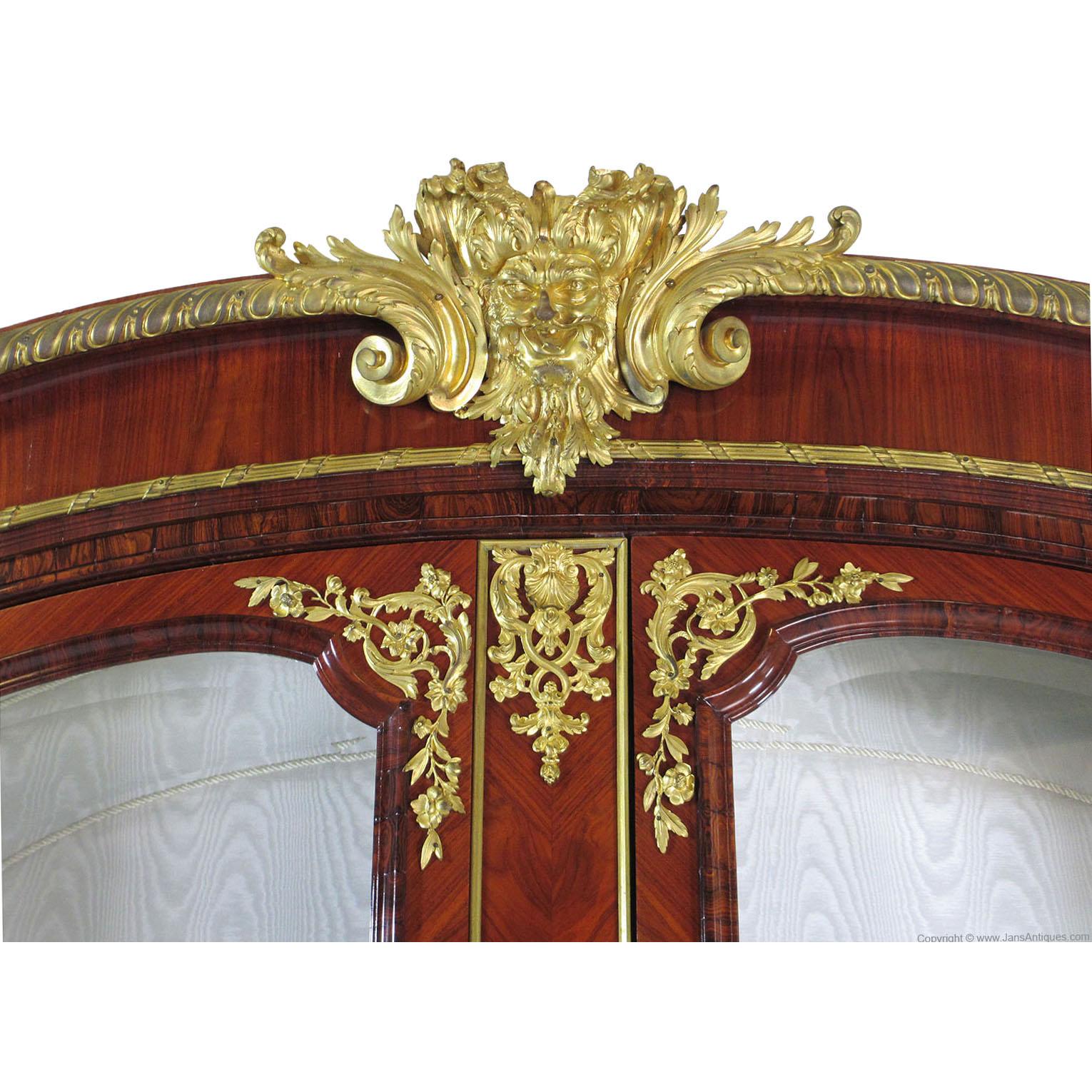 Très belle vitrine à deux portes de style Louis XIV-XV en bois de roi et orfèvrerie, le dessus arqué est surmonté de moulures et de garnitures en bronze doré, L'intérieur est tapissé d'un tissu moiré crème, équipé de trois étagères en verre et d'un