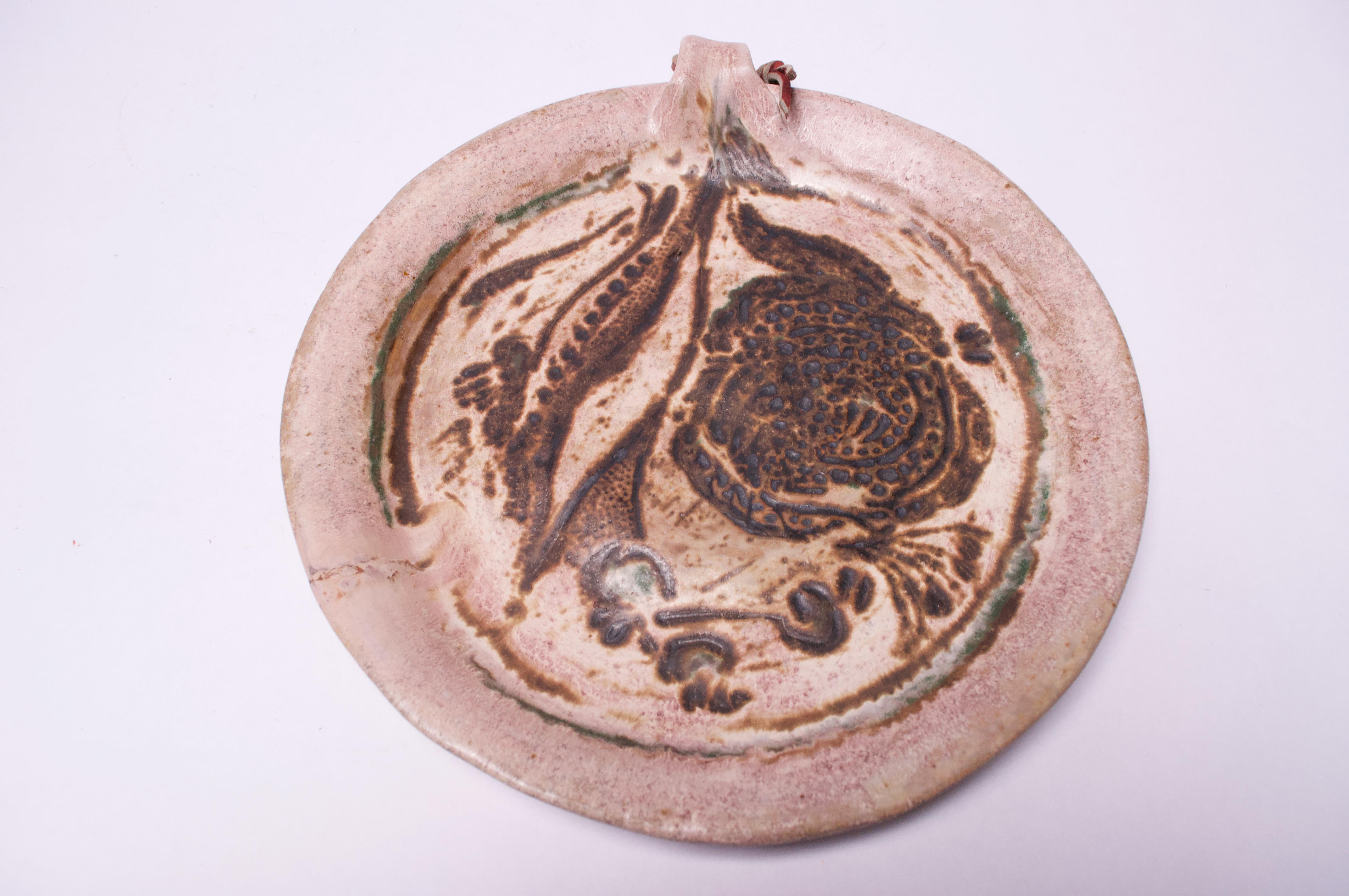 Attractive stoneware charger / decorative plate in muted palette of pale pink and brown with pops of green and a strip of red leather attached to the handle. Nice textural elements present (the floral design is composed largely of small dots /