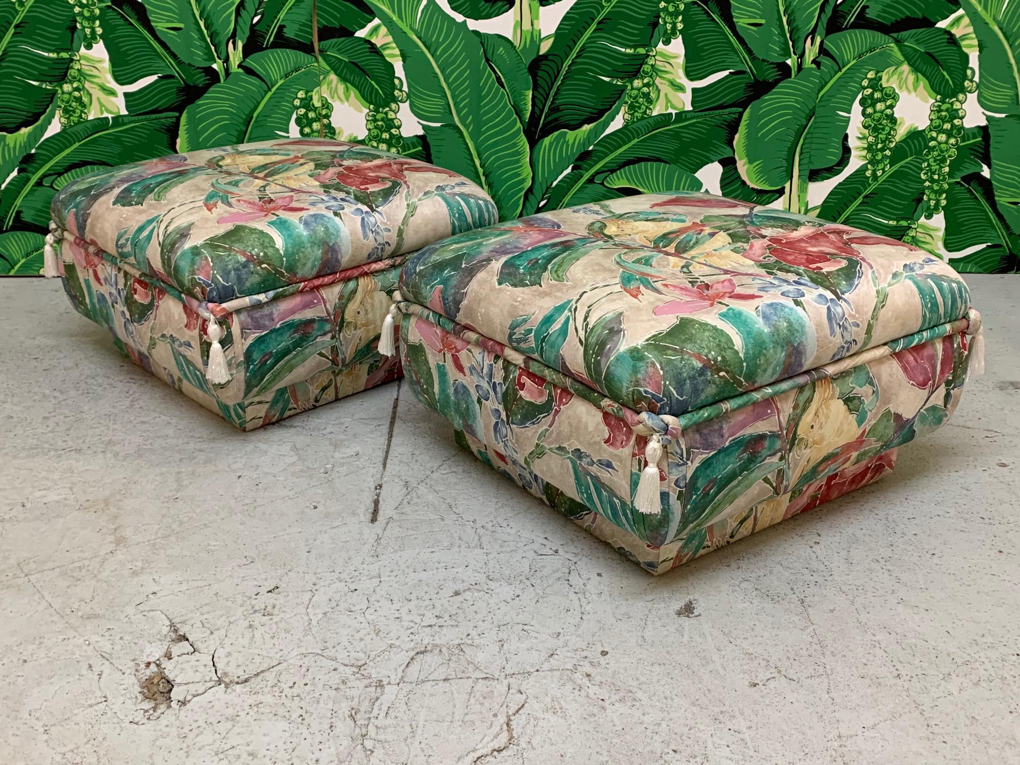 Matching pair of ottomans feature a bold print with tropical birds and flowers along with decorative tassels and an upholstered plinth base. Can be used as seating or footstools. Very comfortable! Very good condition with no stains, tears, or marks.