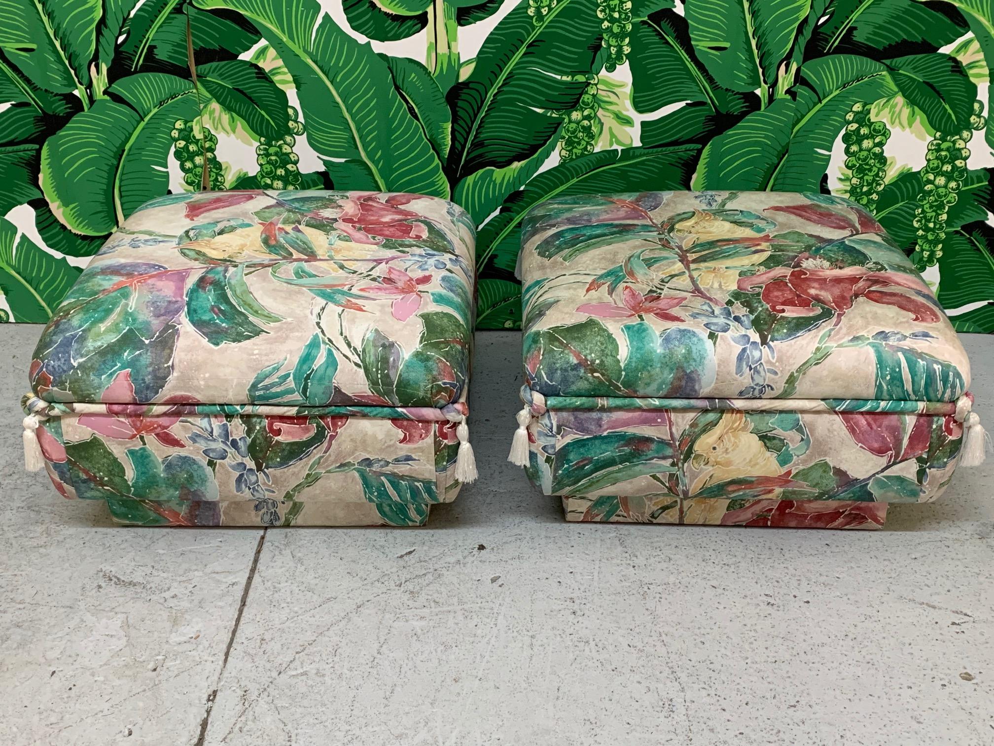 Matching pair of ottomans feature a bold print with tropical birds and flowers along with decorative tassels and an upholstered plinth base. Can be used as seating or footstools. Very comfortable! Very good condition with no stains, tears, or marks.