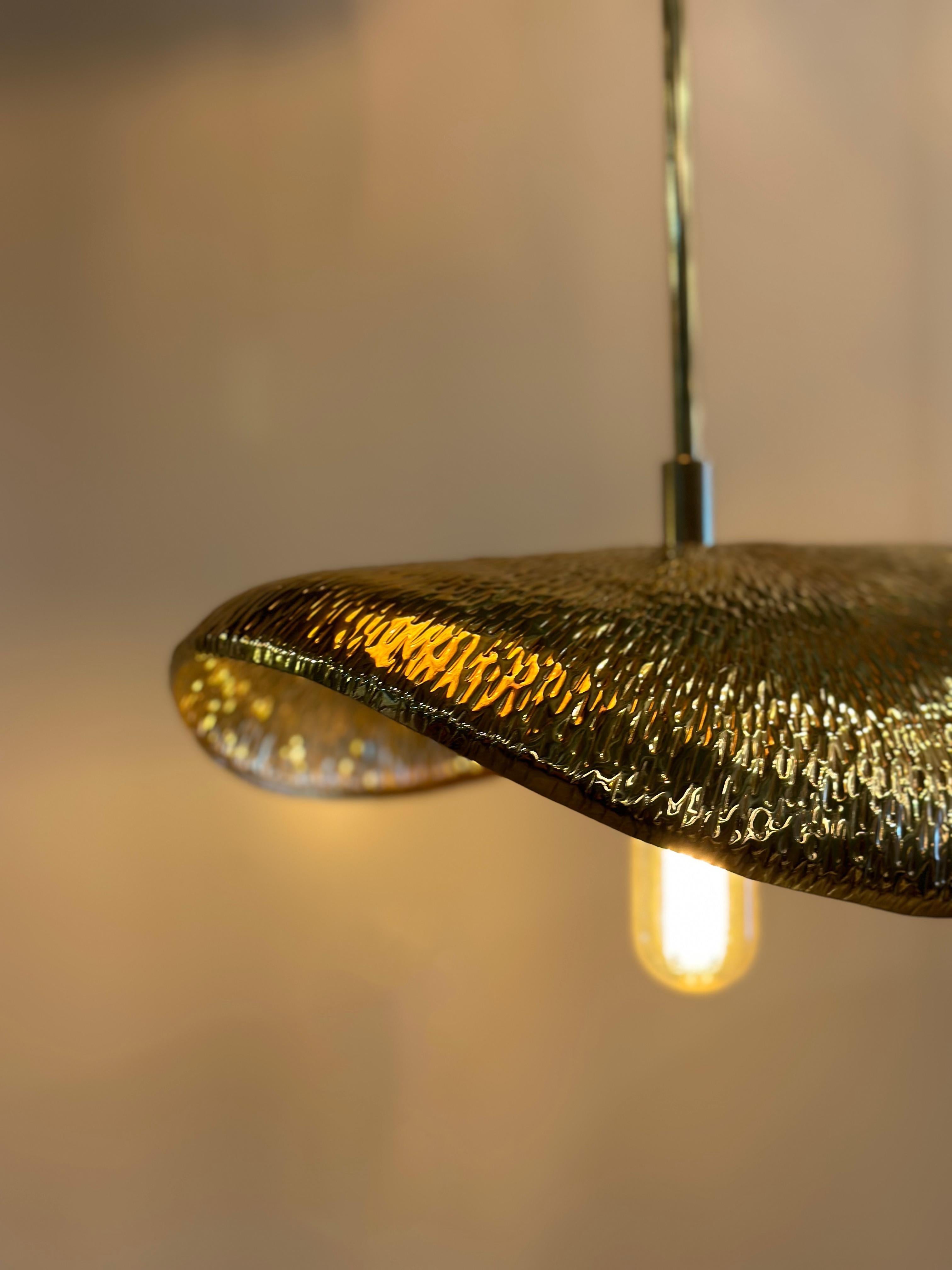 PALMA Medium Brass Hammered Pendant, a masterpiece that seamlessly marries the elegance of nature with meticulous artisan craftsmanship. Crafted from a single layer of brass meticulously hammered into a captivating form, this pendant reflects the