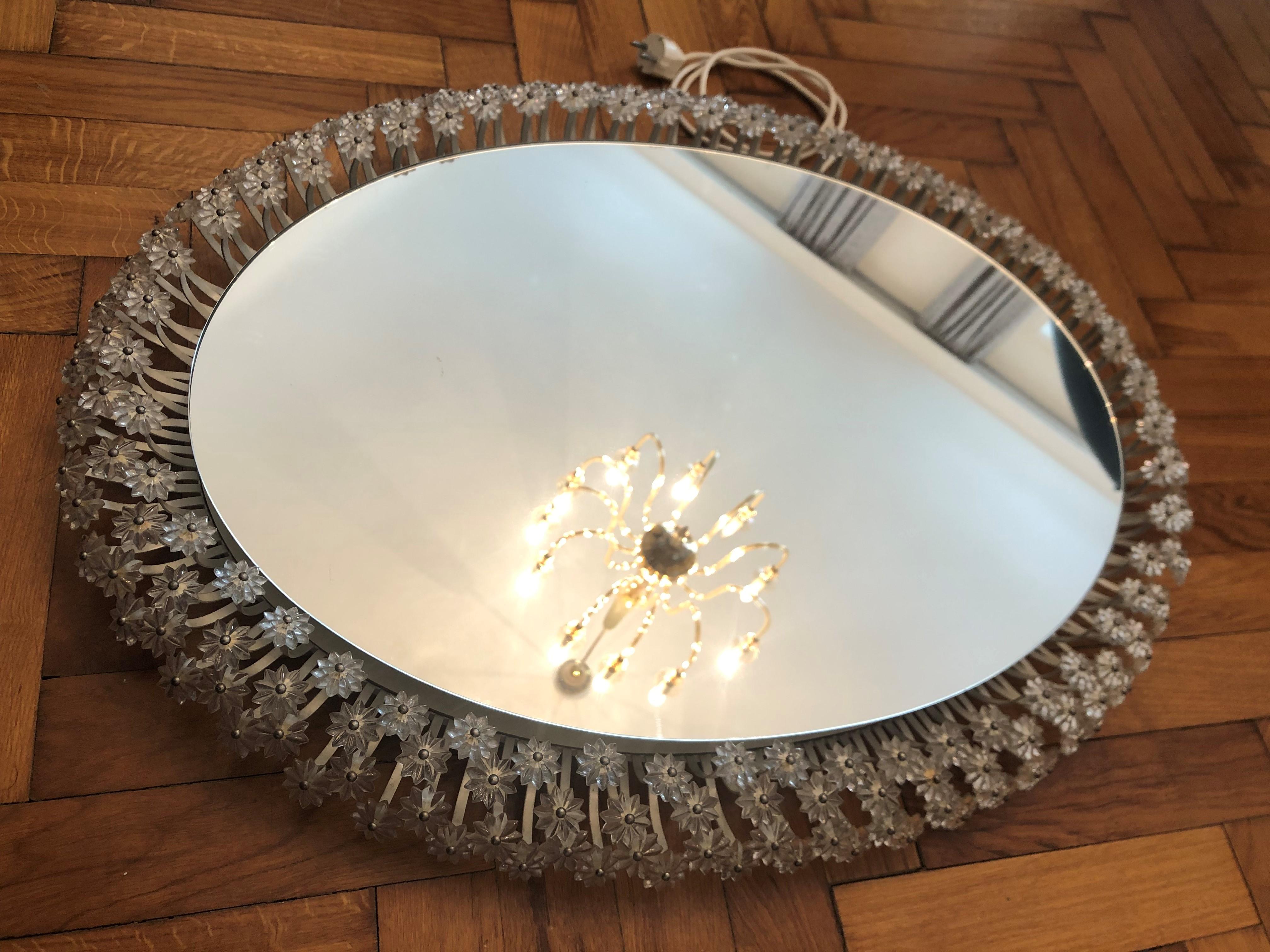 Mid-20th century illuminated oval wall mirror by German manufacturer Palwa. 
The mirror is framed with a border of 4 ranks of flowers and petals made from faceted real glass star shaped crystals and beads that sparkle in the light. 
The frame is