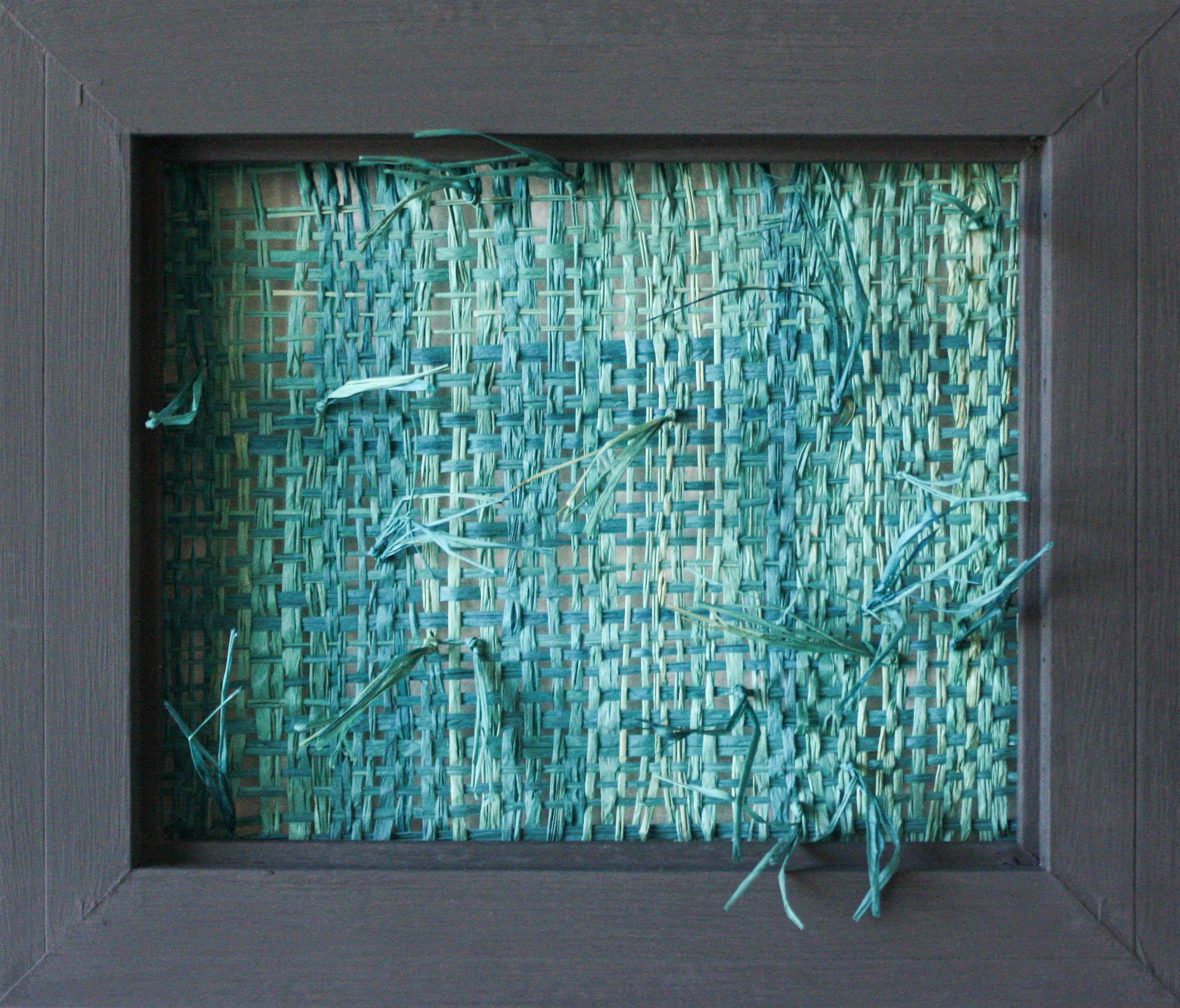 This woven sculptural wall hanging is created using blue raffia mounted inside a wooden frame. Its organic nature is complemented by a few loose pieces that break out of the rectangular scope of the frame.