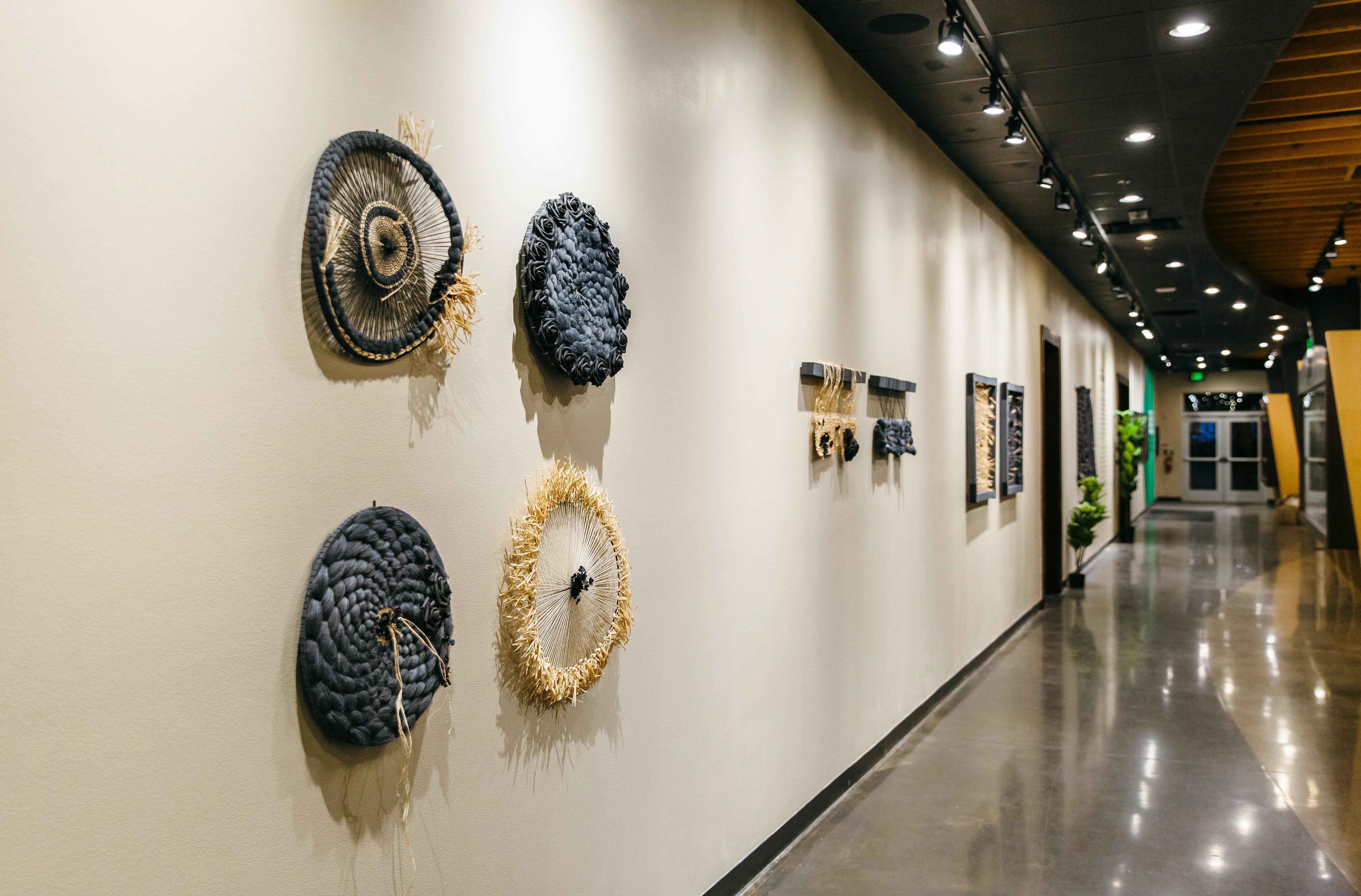 This circular woven sculptural wall hanging is created using raffia, cotton thread, artificial flowers, and latex paint on a wooden hoop. It is beige with black floral accents. The organic mixed media materials display beautifully on the wall.