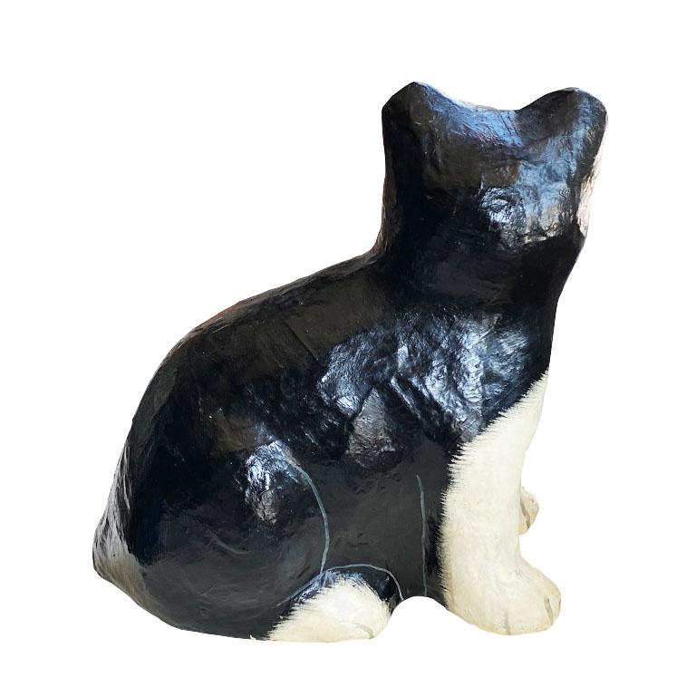 A black and white papier mâché kitten sculpture. This fun figurine depicts a cat with white paws and nose, and a black body. Its soft black eyes seem to look right at you! This would be a fun piece to style on a bookshelf, or as a gift for a feline