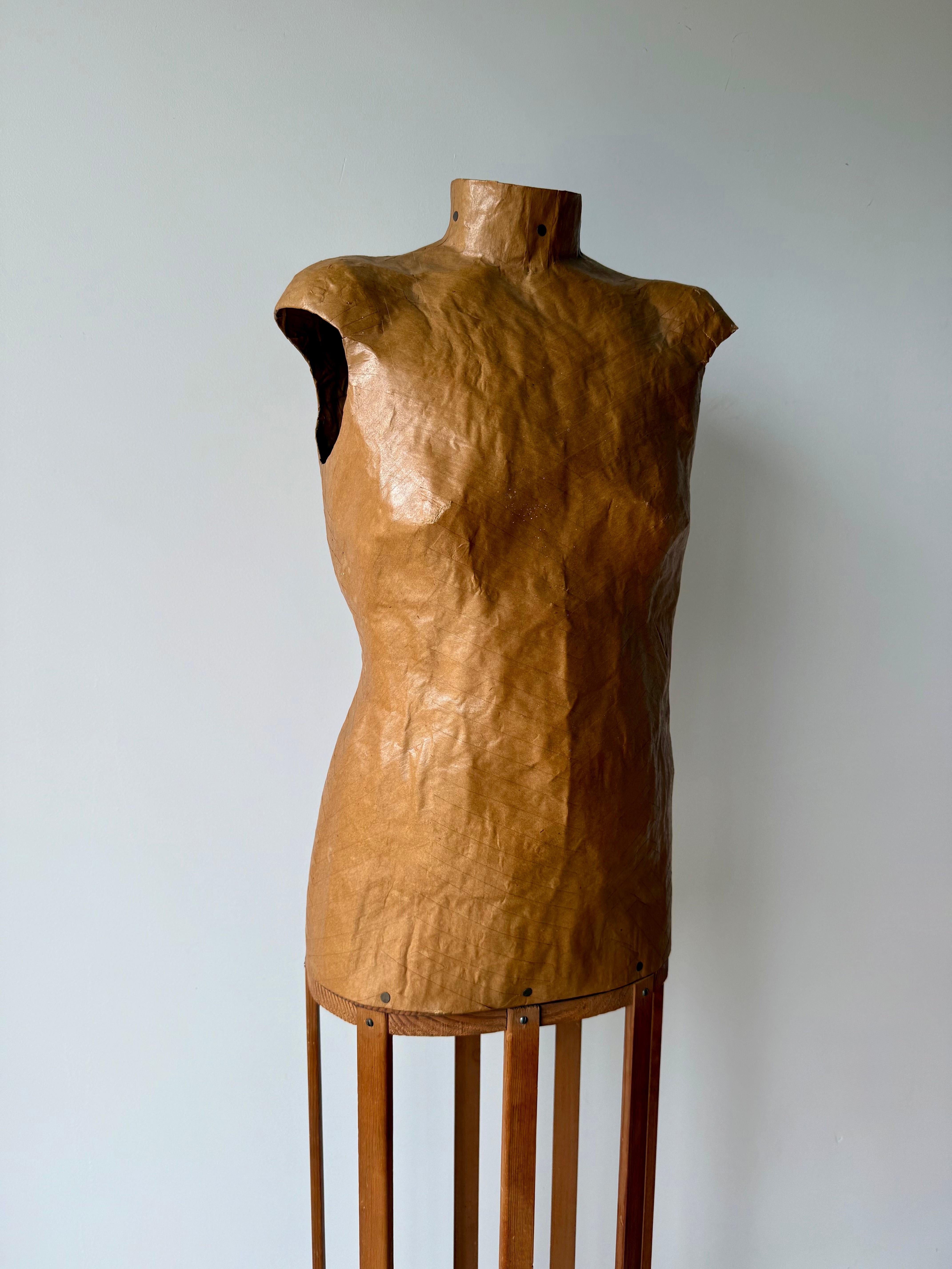 A sculptural papier-mâché dress form mounted on its original freestanding wooden slat stand, dating from the mid-20th century. The form is lightweight and crafted from layered brown paper, hand-shaped and finished with a varnished surface that