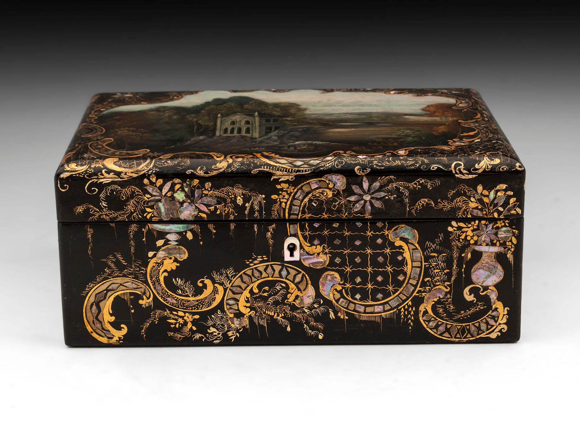 Papier mâché sewing box beautifully painted with a landscape scene, bordered by gold floral designs adorned with inlaid abalone. 

The interior is lined with red silk velvet and features a removable tray which houses various sewing tools including
