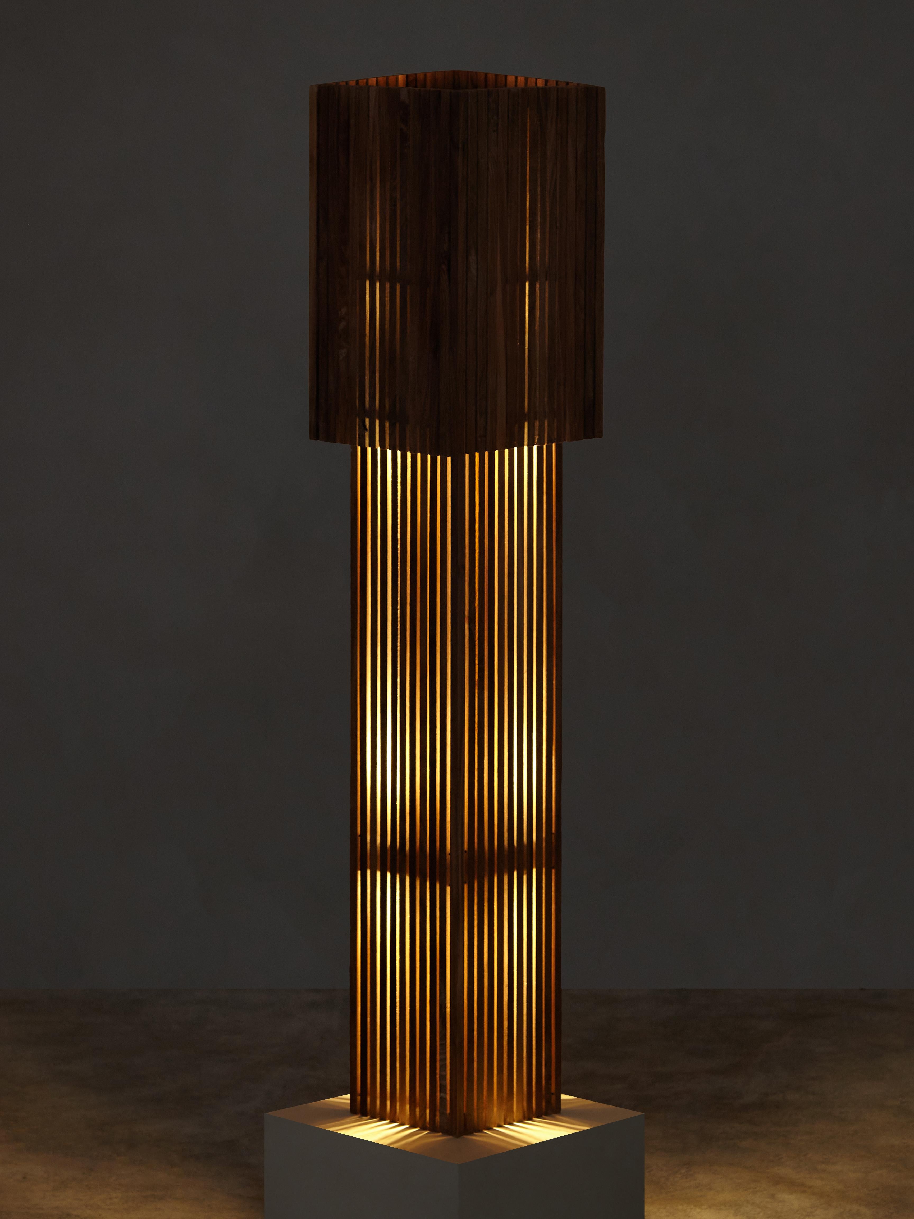 Des chevilles en bois brossé à la main bloquent et dirigent l'ampoule LED dissimulée de la lampe, projetant une lueur inégale à travers les bandes verticales du corps de la lampe. L'abat-jour amovible fonctionne comme un variateur manuel, permettant