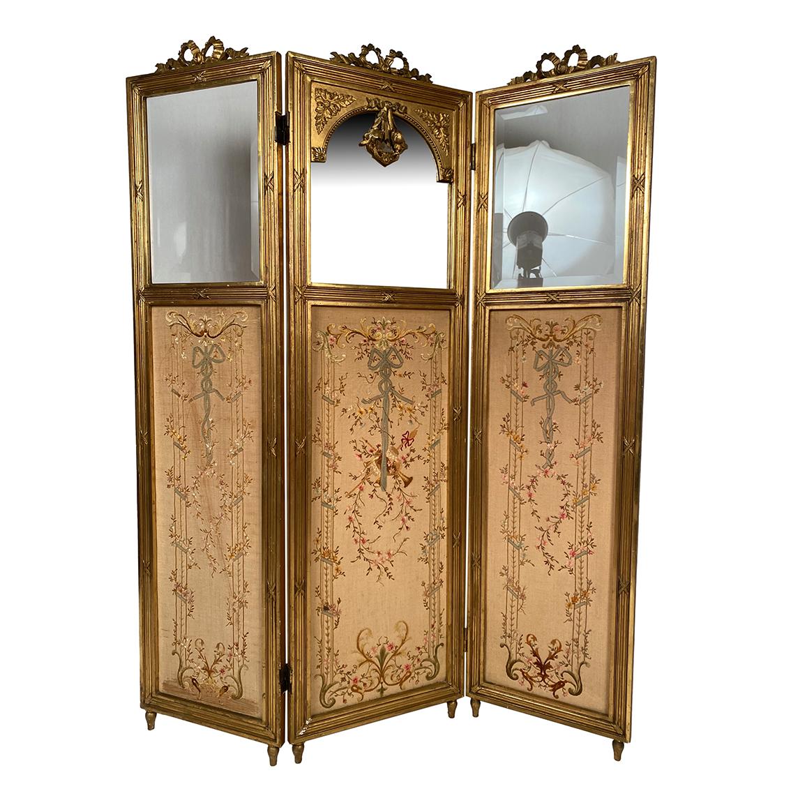 Paravent en bois doré de style Louis XVI, vers 1900
Trois feuilles
Tissu finement brodé en partie basse,
miroir en partie haute centrale et verres biseautés en partie haute pour les feuilles latérales.
Chaque feuille sommée d’un nœud de ruban