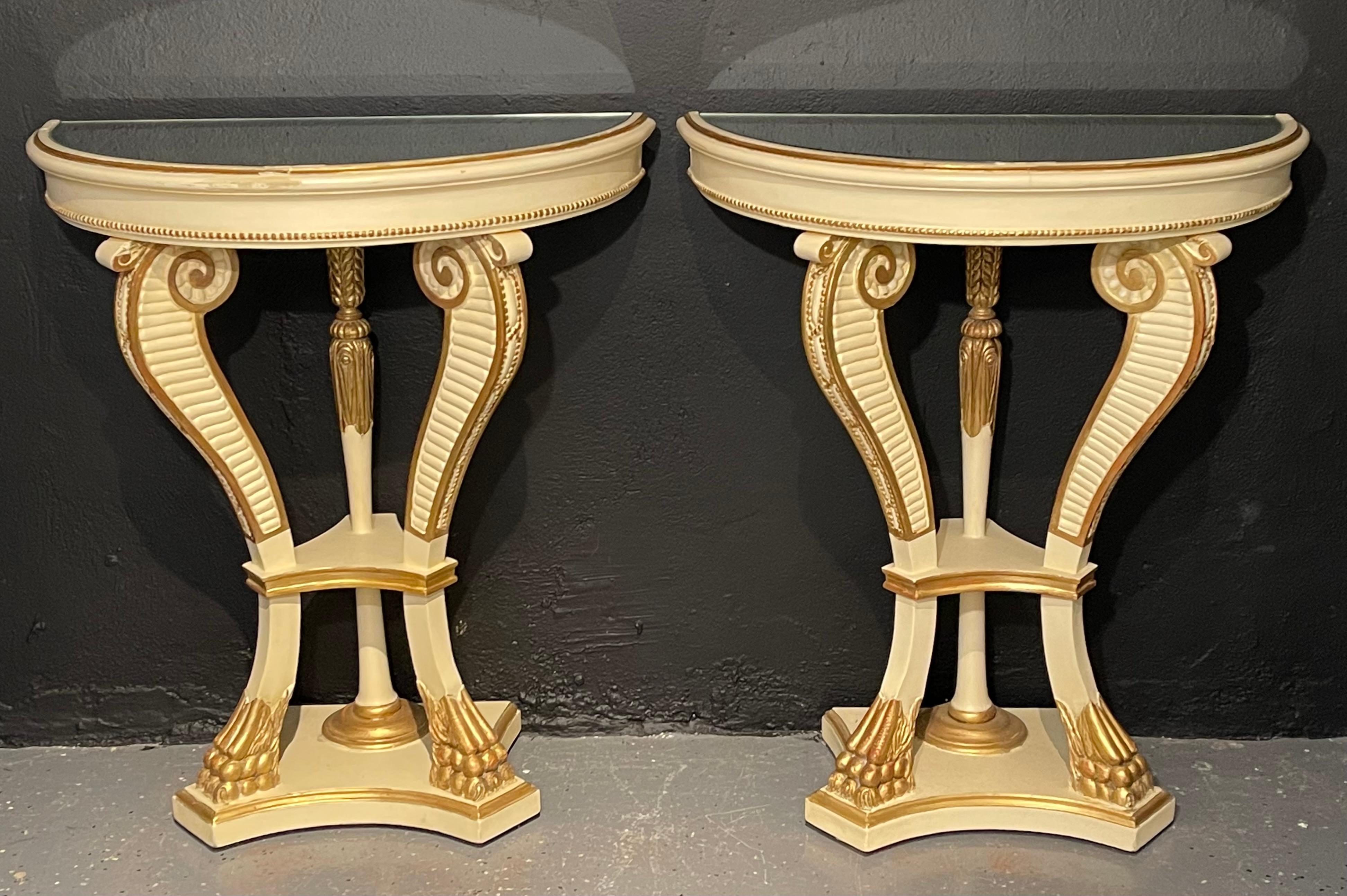 A pair of parcel paint and gilt decorated mirror top demilune console or sofa tables with claw feet. Each having a linen white painted background with a gilt gold design having a black marble top in the demilune form. Supported by scroll reeded