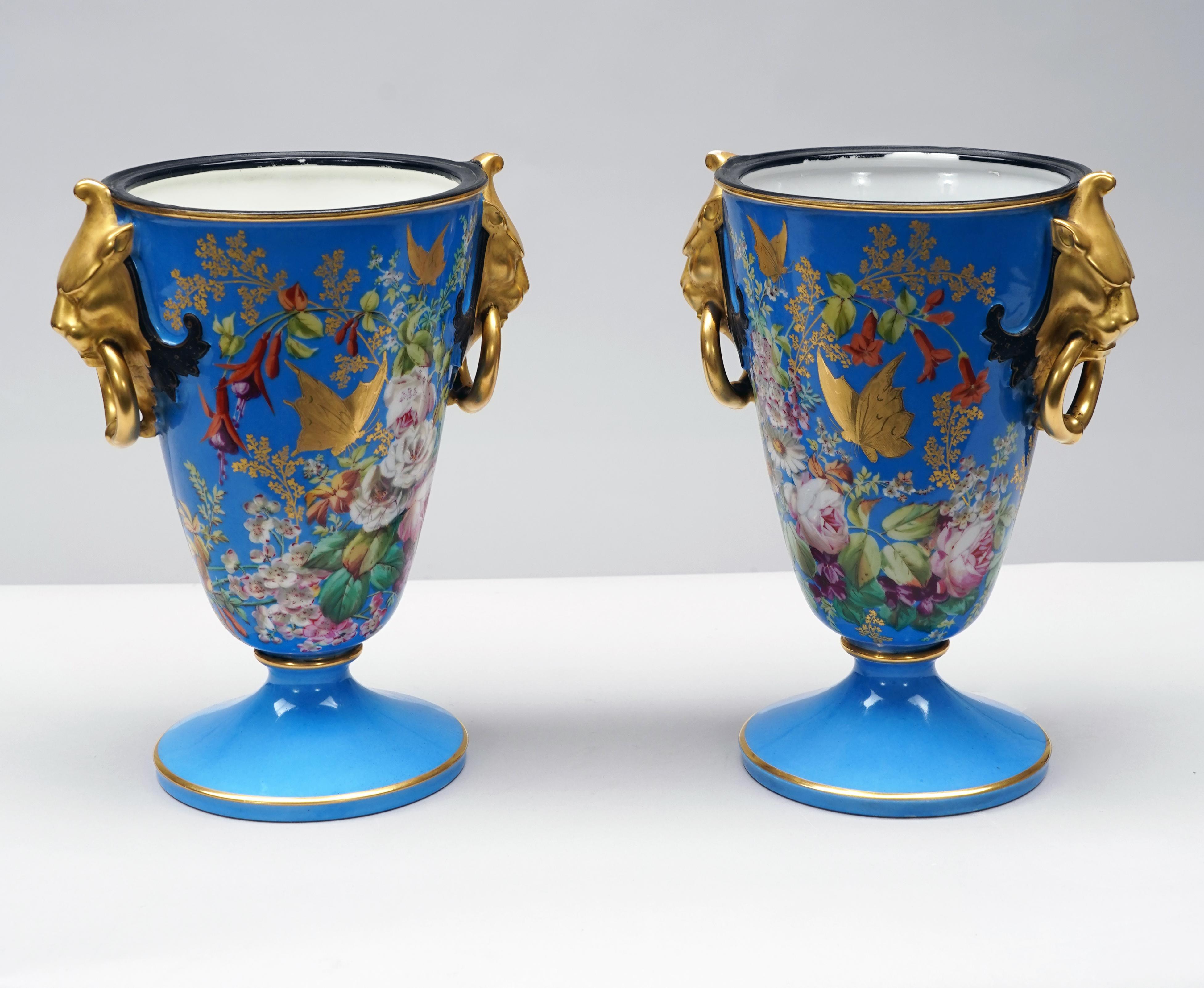 Bellissima coppia di vasi in porcellana bianca riccamente decorati con splendide composizioni floreali policrome e farfalle impreziosite dall'oro su sfondo blu, incorniciati da manici in porcellana dorata a forma di testa di leone che racchiudono un