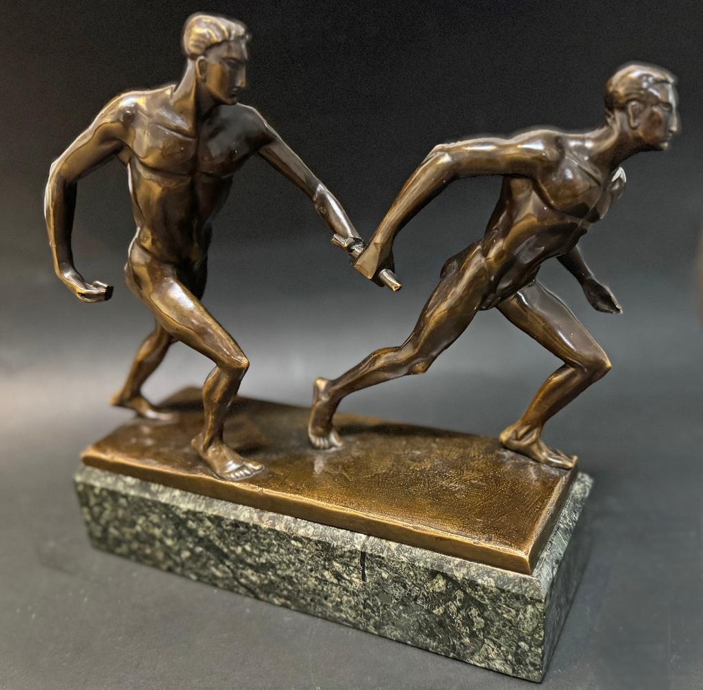 Con sus físicos cincelados y sus rostros angulosos, esta escultura de bronce de dos figuras masculinas desnudas pasándose el testigo en plena carrera de relevos es el epítome del arte Art Déco estilizado.  Los dos hombres están muy centrados y