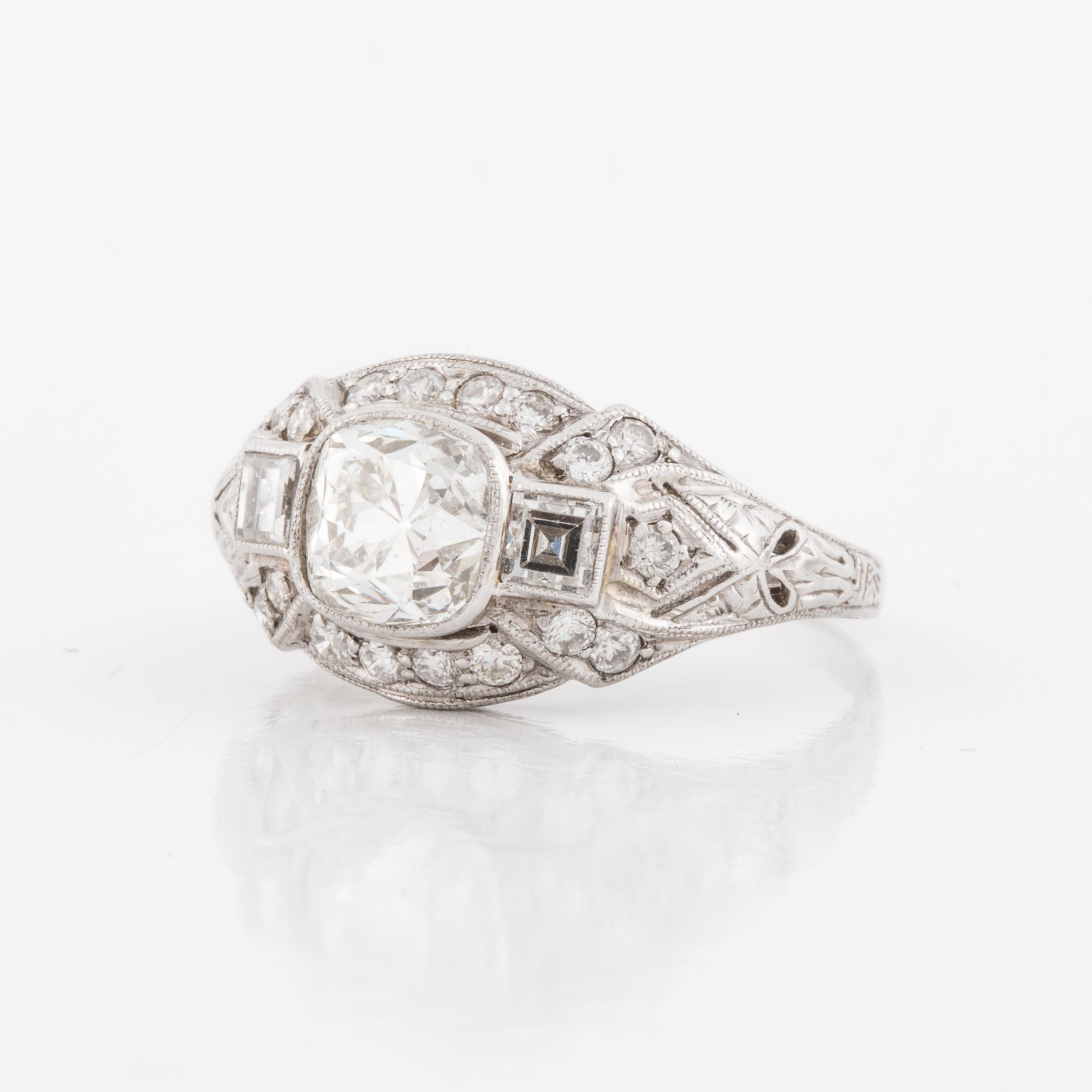 Art Deco platinum ring consisting of a central bezel-set antique cushion-cut diamond flanked by square step-cut diamonds in a finely-detailed openwork mounting enhanced by bead-set diamonds and engraving.  Ring features one central antique