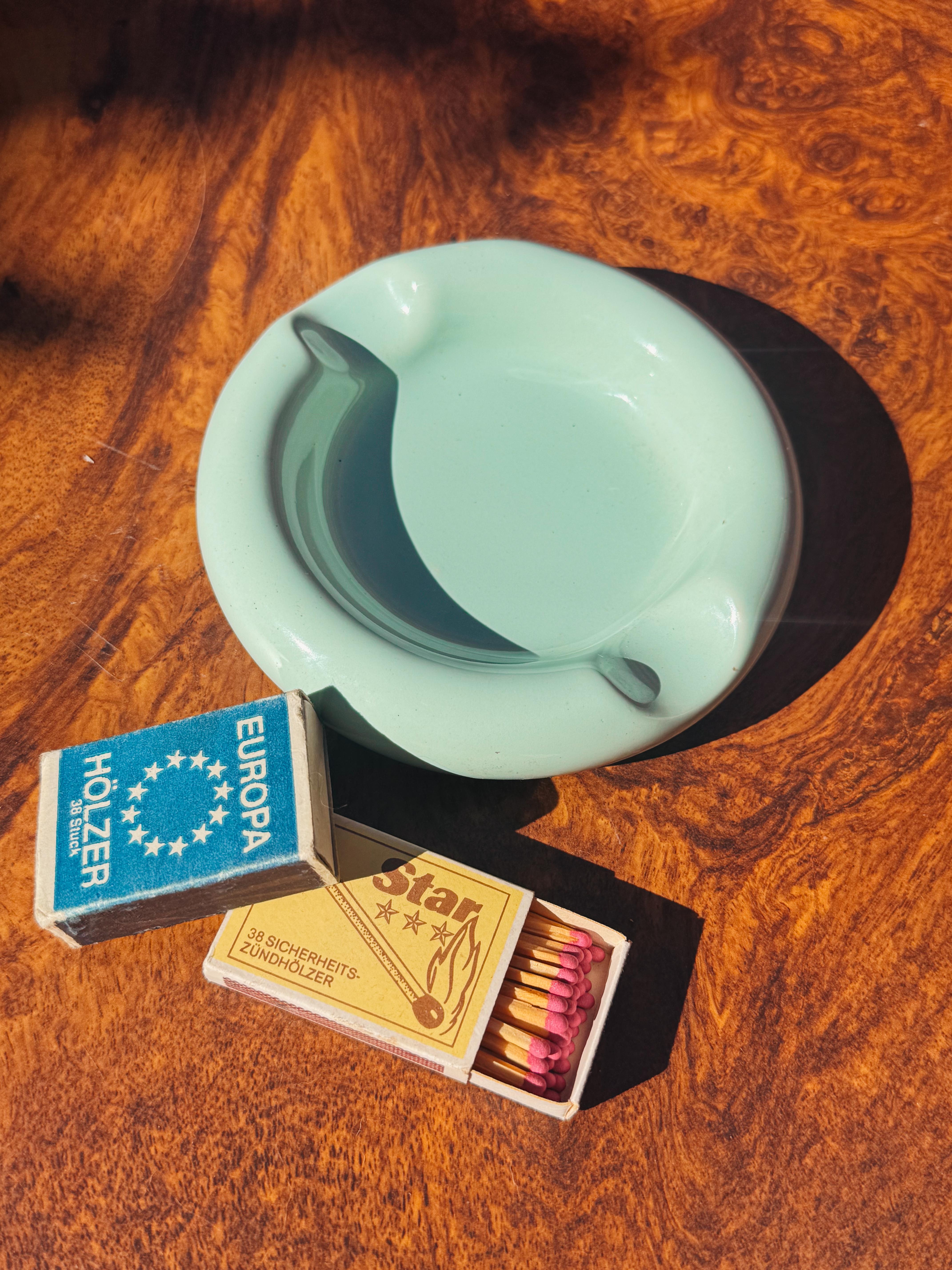 Un elegante cenicero de cerámica verde pastel de los años 80, que refleja la estética lúdica pero funcional del diseño posmoderno. La pieza presenta una sencilla forma redonda con tres apoyos, acabada en una suave superficie esmaltada que resalta su