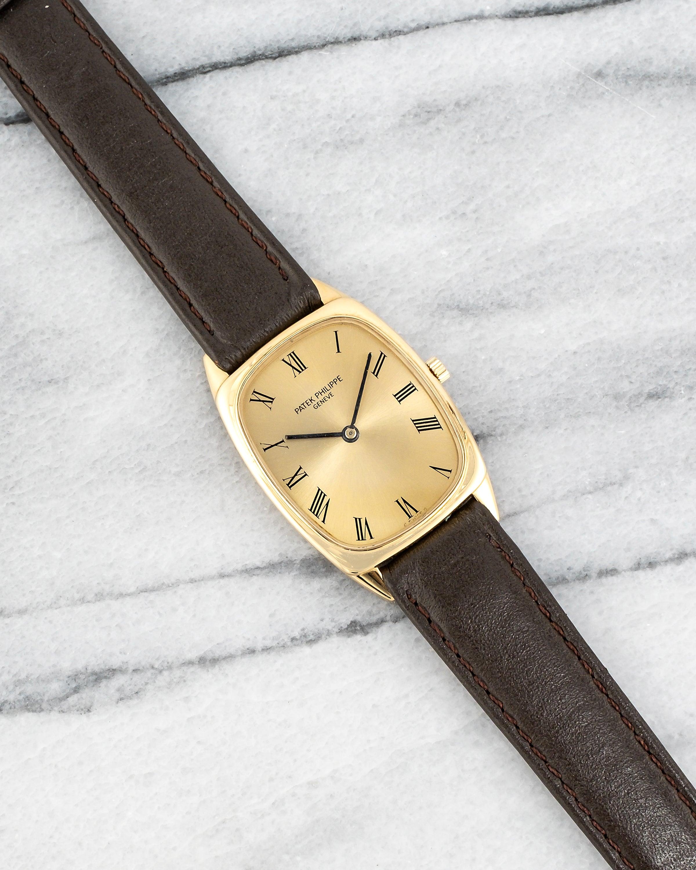 Here's a vintage Patek Philippe Tank manual wind in 18k yellow gold with a black Roman numeral dial from the 1970s. Measurements include a 29mm yellow gold case and 16mm lug width. It's on an aftermarket brown leather strap. Yellow gold case and