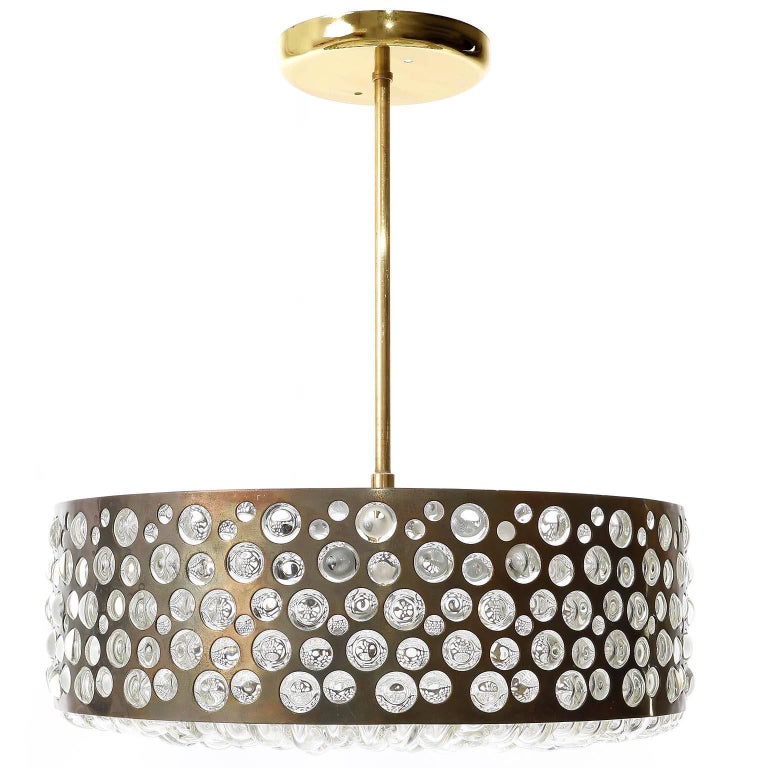 A fantastic disc shaped light fixture by Rupert Nikoll, Vienna, manufactured in midcentury, circa 1950.
The lamp is made of a seamless brass belt with holes of varying sizes. The hot and molten glass was handblown through the belt. The brass got a
