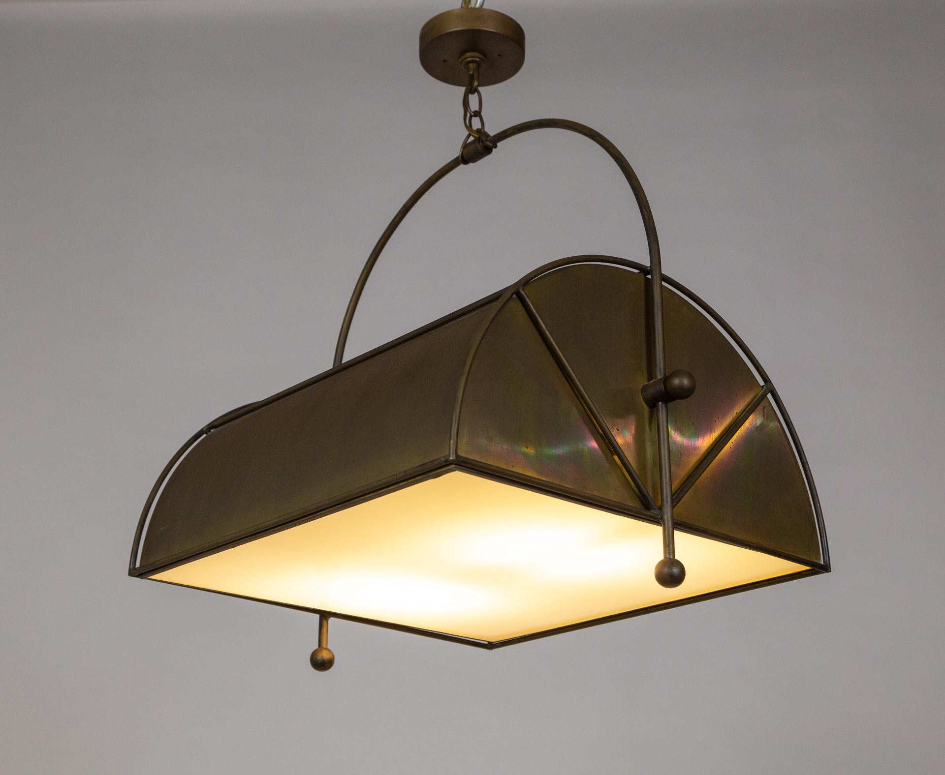 A unique, custom, pendant light; half-cylinder shaped with an arching handle hanging from a short chain and a white glass diffuser on the bottom. Constructed with matte patinated brass by Morrison Custom Lighting. Perfect for over an island, dining