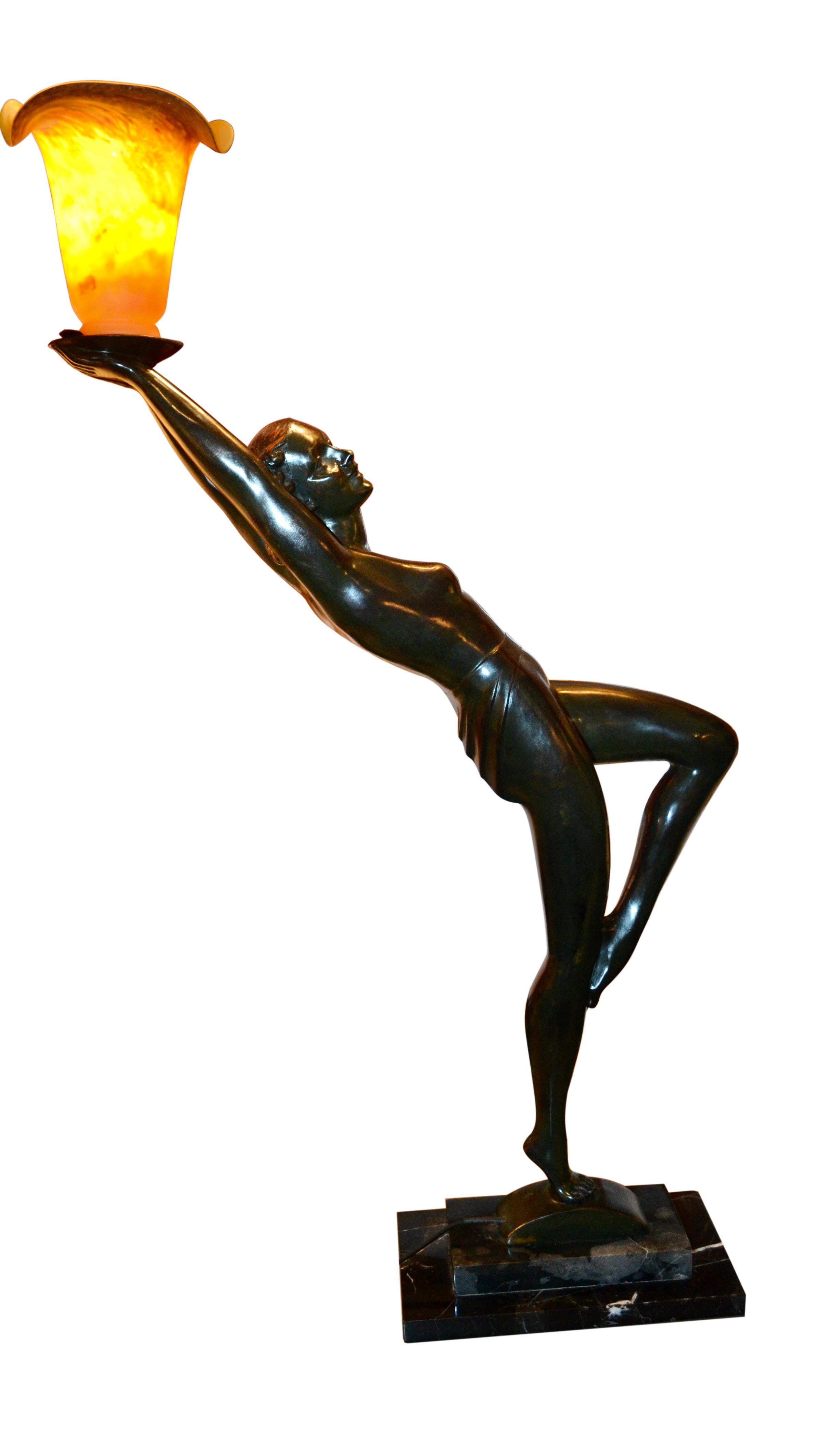 A patinated bronze figure of a stylized Art Deco nude woman shown in a  dance pose. She stands on her right leg, left leg up and angled; back arched and arms aloft holding a colored glass tulip shade. The bronze is mounted on a stepped dark green