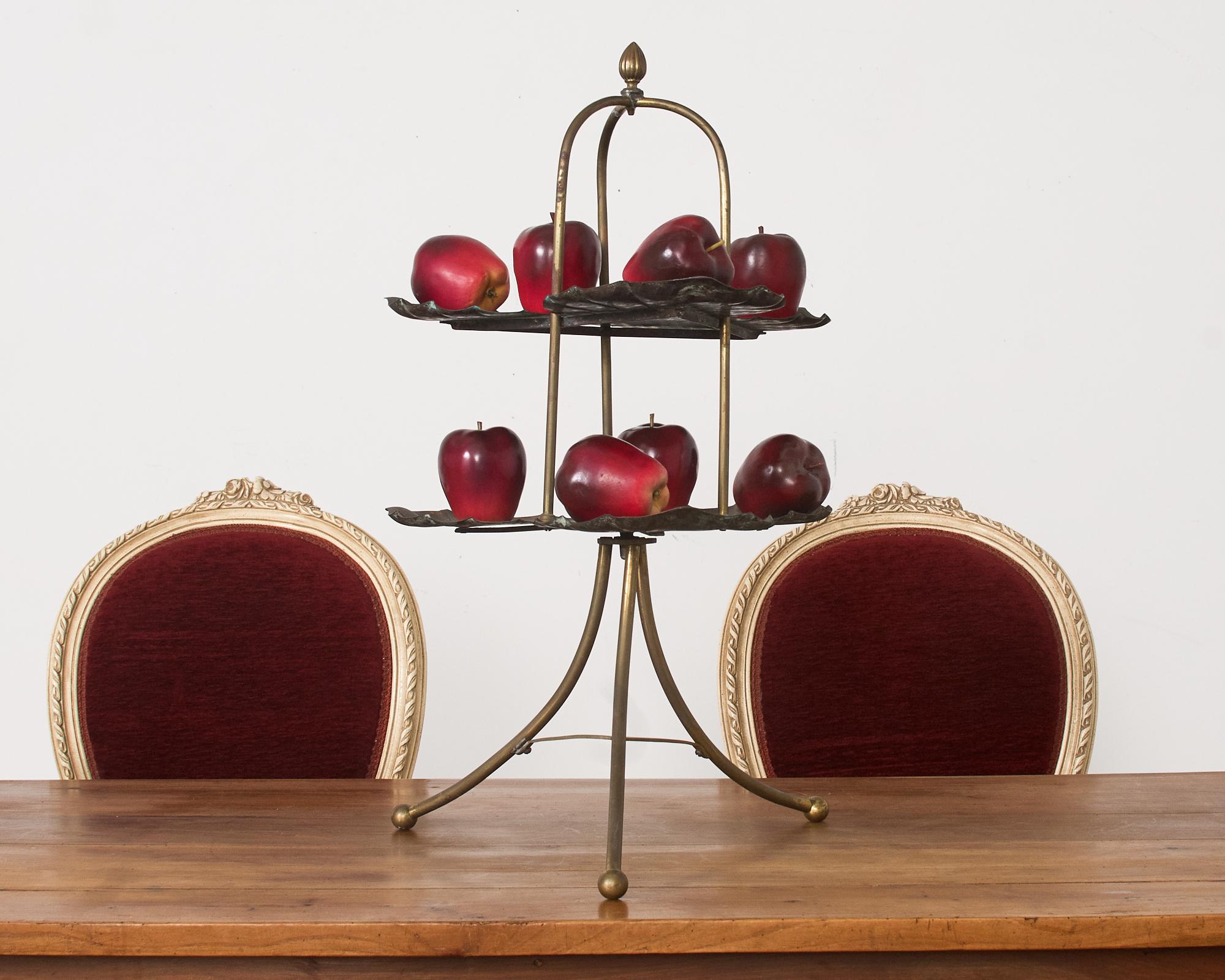 Beautifully patinated bronze and brass tiered dessert or muffin stand featuring lotus leaf or water lily pad tiers. The stand has a fluted finial on top with three legs conjoined to the two tiers. The top is supported by a tripod base ending with