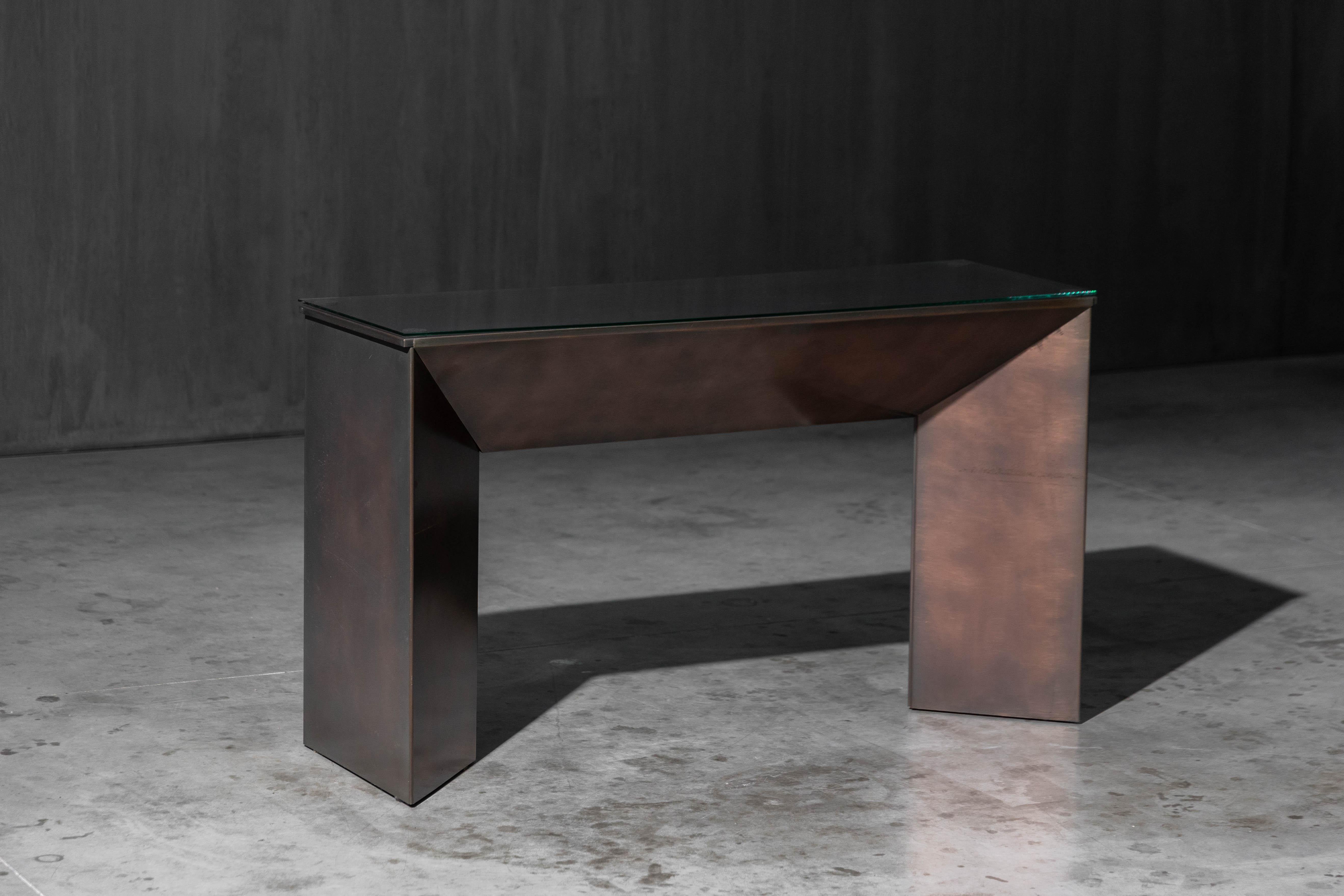 Tavolo consolle della casa di design belga Belgochrom, caratterizzato da una base minimalista in metallo patinato bronzo e da un elegante piano in vetro. I toni caldi e ricchi del bronzo creano un sofisticato contrasto con la fredda trasparenza del