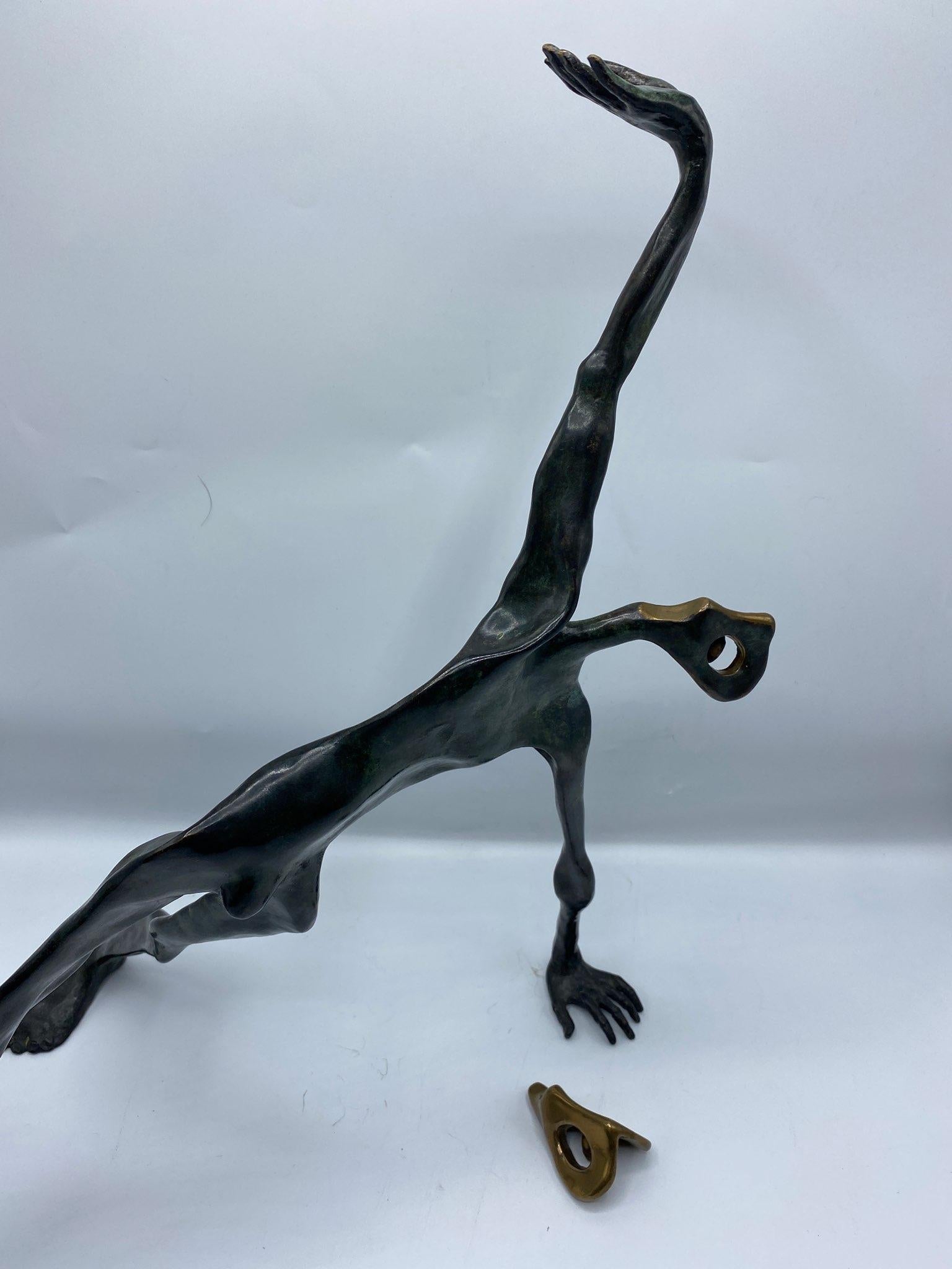 Bronze figure of a male nude and a small bronze mask. The figure has his legs stretched with one hand to the ground and the other to the sky. The small bronze cast mask is placed separately next to him. The bronze of the man is patinated in a deep