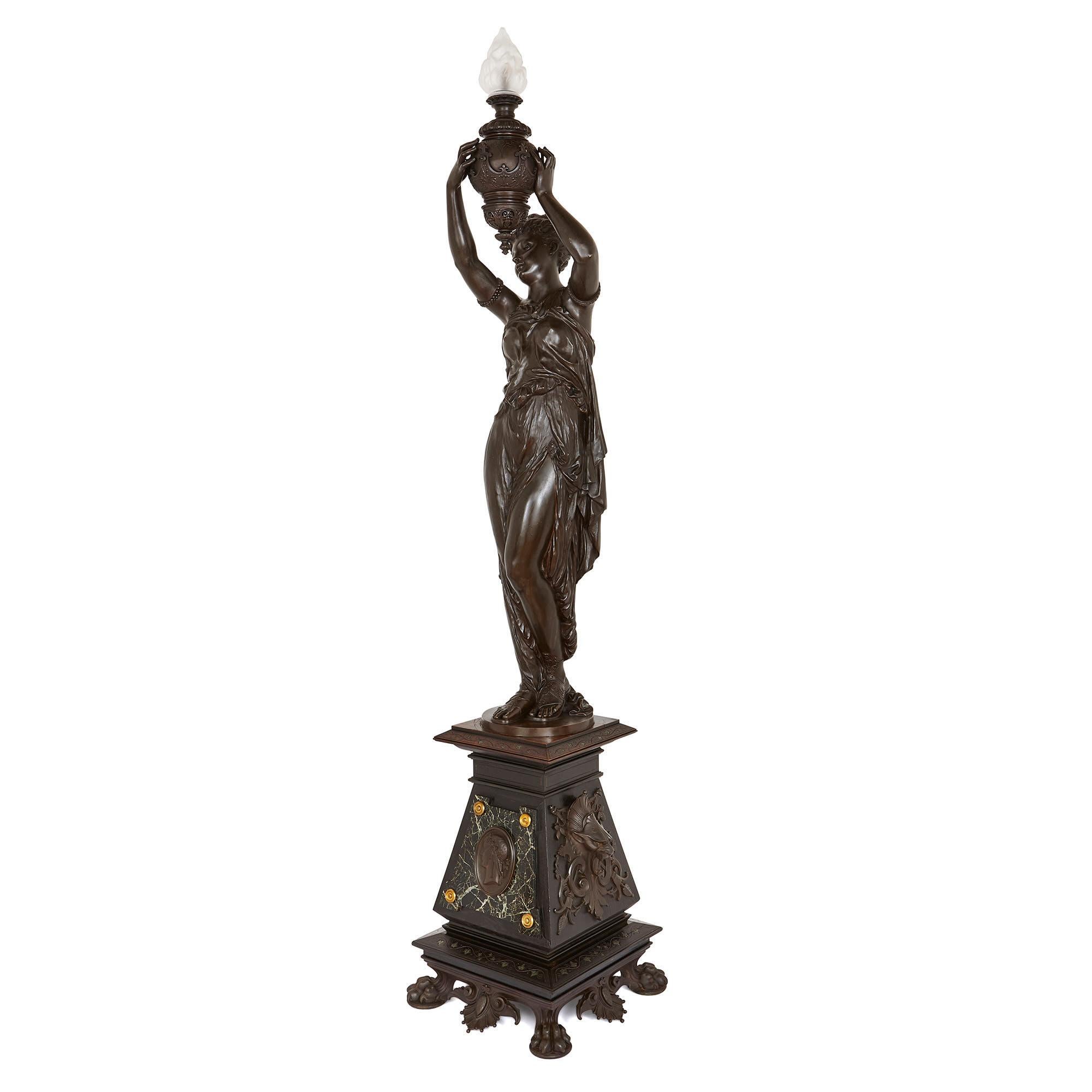 This beautiful piece consists of a patinated bronze female figure holding an urn with a glass flame, which serves as a lamp, all standing on a tall and finely decorated wooden plinth. 

The plinth is crafted from ebonized wood, set on a slim