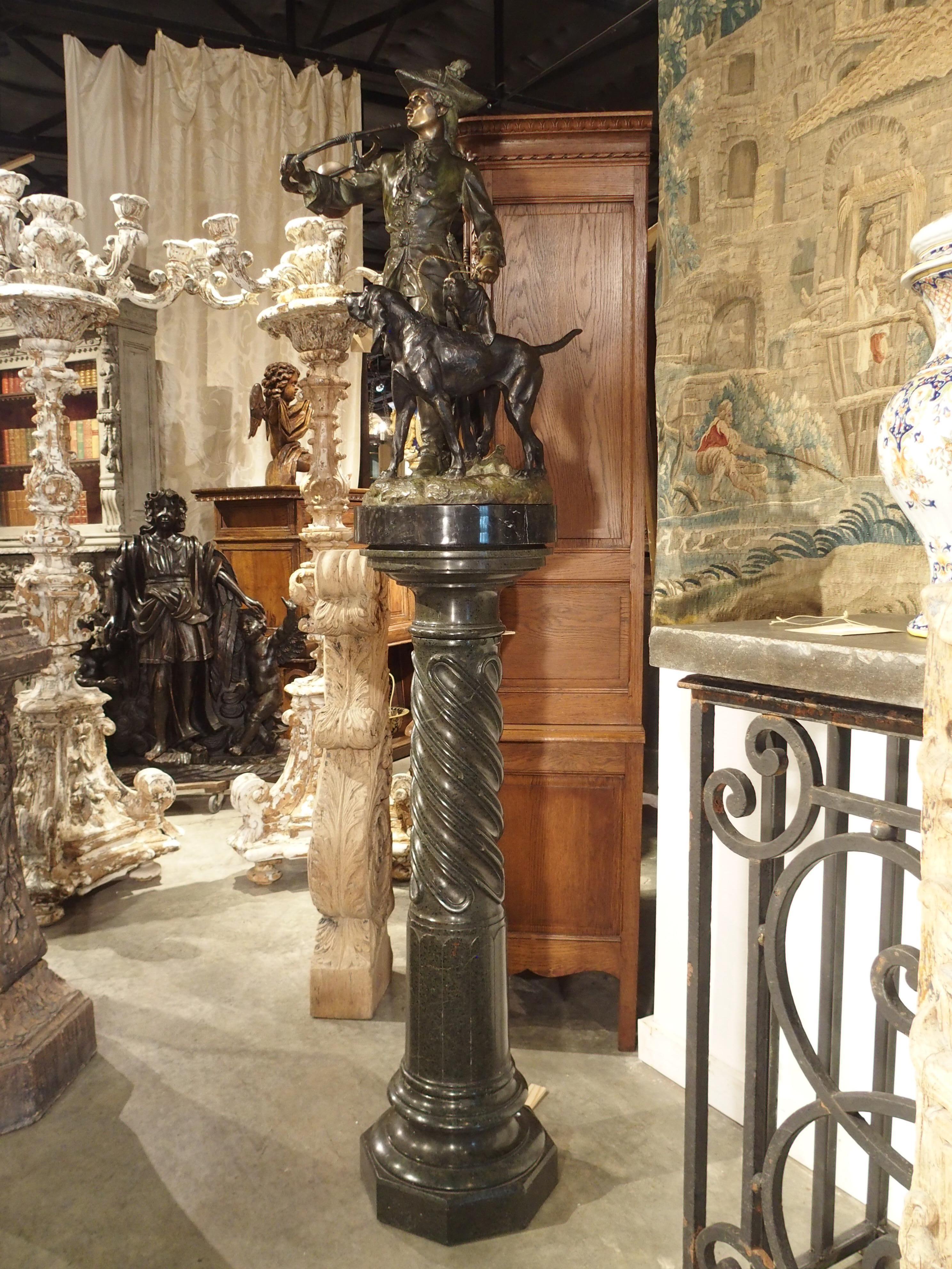 Cette statue en bronze patiné représentant un chasseur avec des chiens courants repose sur un haut piédestal en marbre. La statue très détaillée est un mélange de bronze naturel, de vert panaché et de patine noire. Un chasseur est un membre de