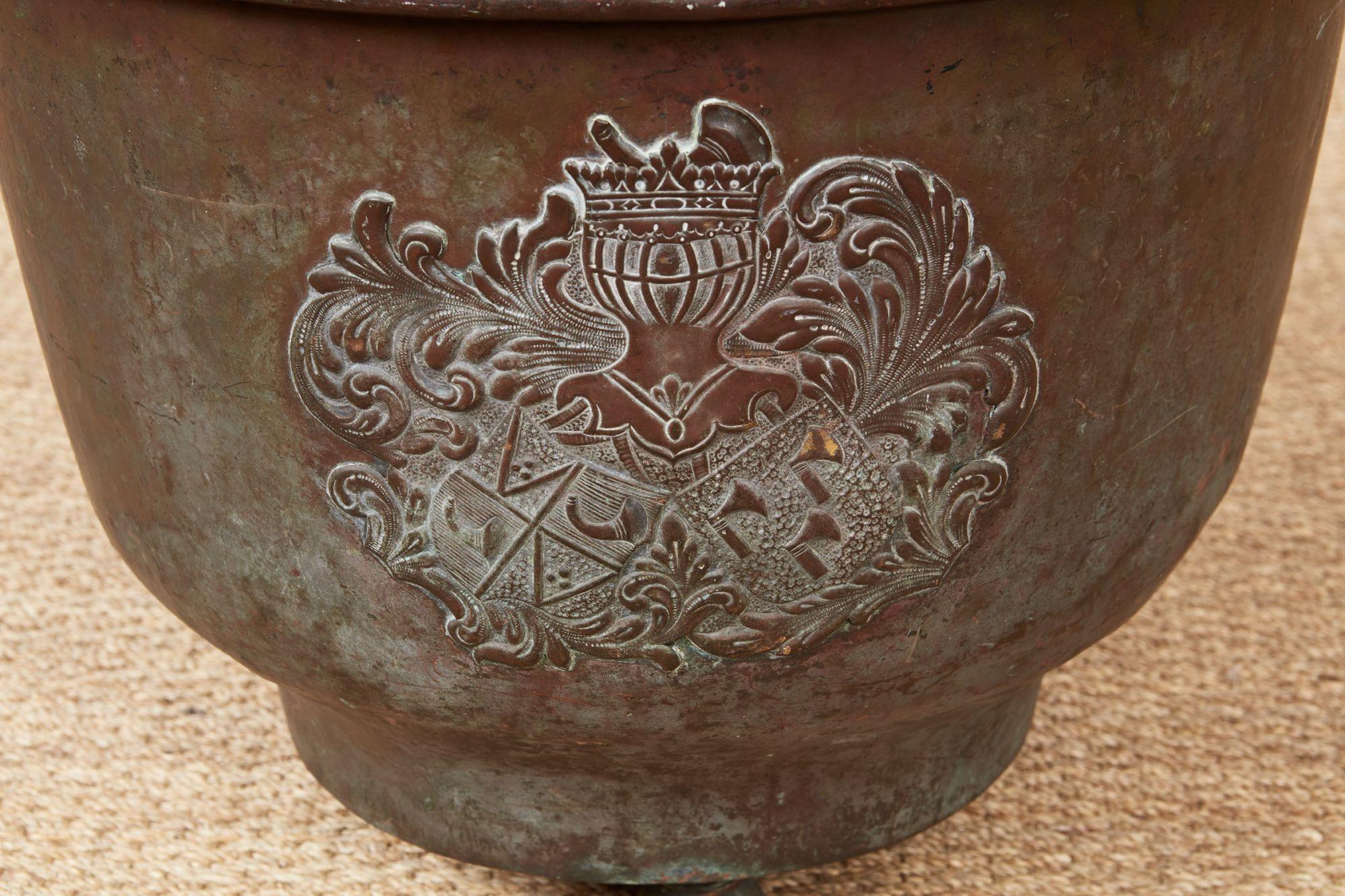 A copper bin of footed bowl form possessing a wonderful bronze patinated surface and featuring a baronial crest with a crown and crests of horns, dividers and axe heads, supported on three bronze ball feet.  Would make a lovely kindling log bin or
