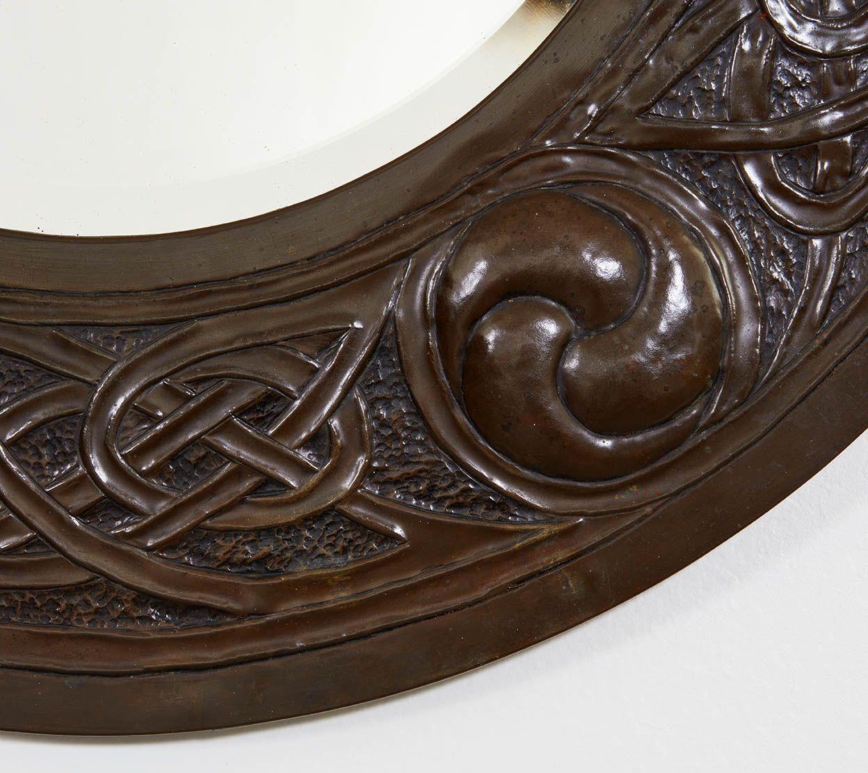 A large round Arts and Crafts copper cushion mirror having raised repousee designs of curved geometric celtic knots between roundels known as triskeles, in a wonderfully rich patinated finish. Retaining original beveled glass.
