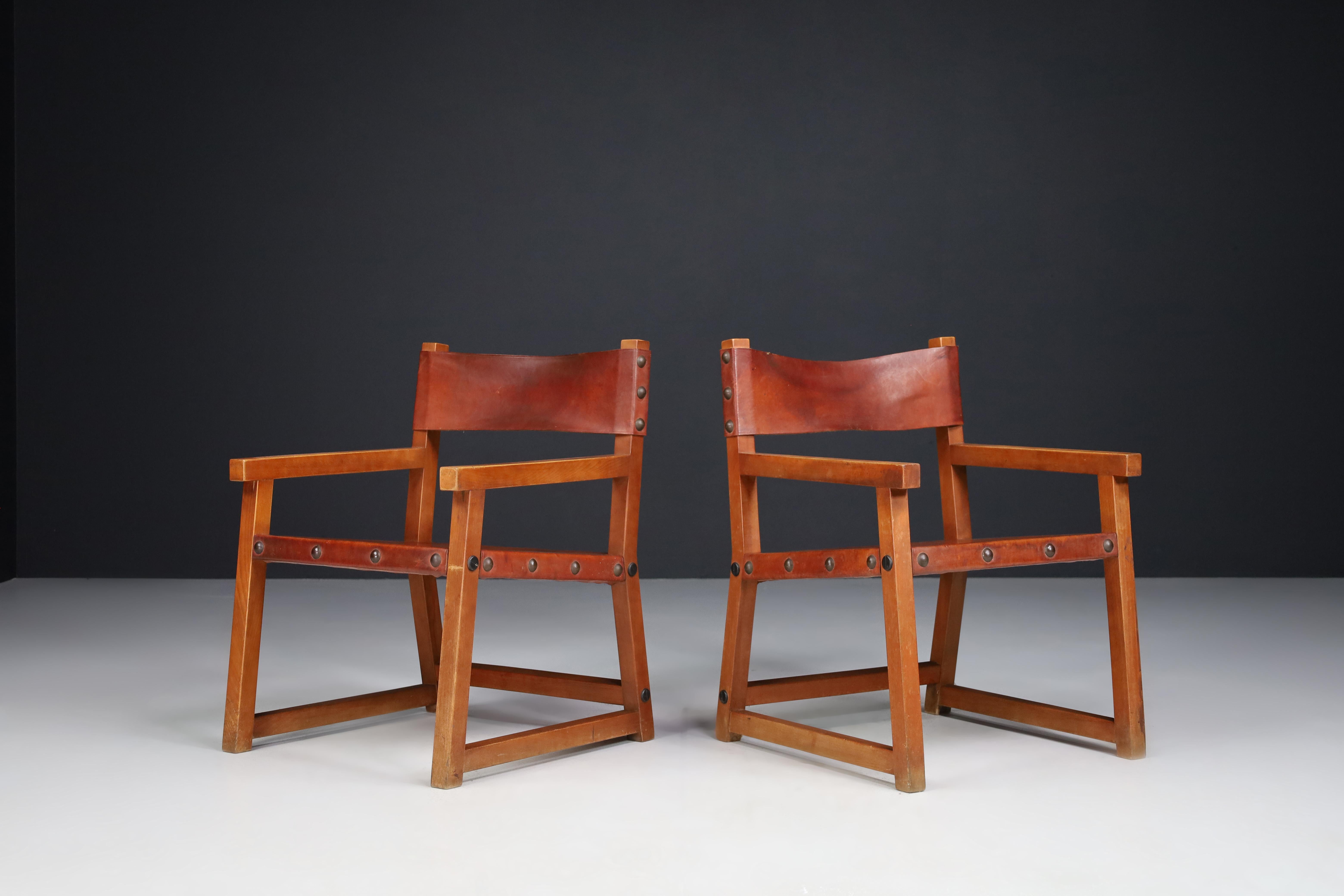 Patinated saddle leather armchairs, Spain 1950s. 

A remarkable pair of Spanish armchairs in solid wood dating from the 1950s with original saddle leather seats, backrest, and domed metal studs. These particular chairs are as solid and stable as