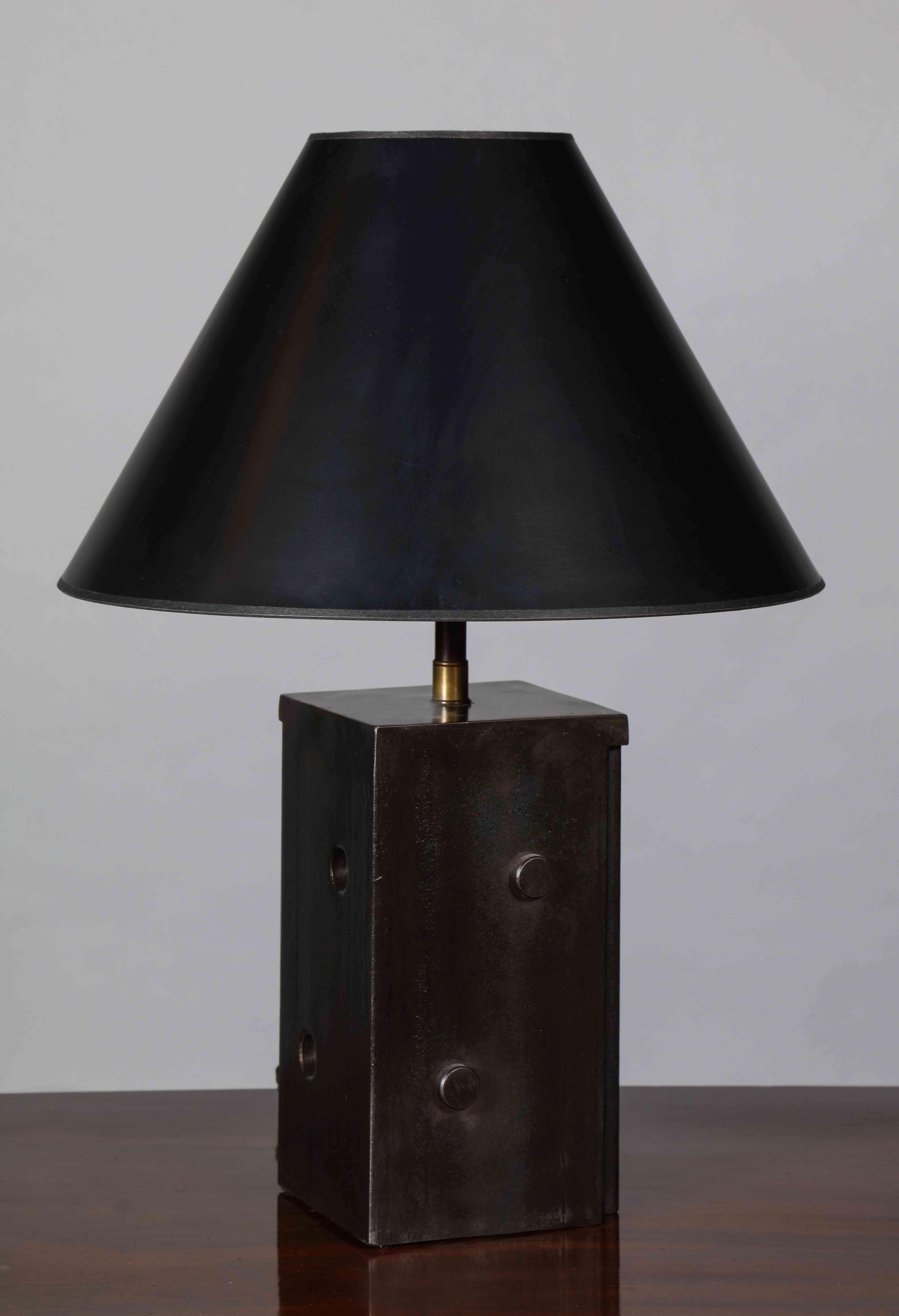 Une lampe de table effilée construite en acier laminé à chaud avec une surface à la fois texturée et polie, avec des découpes circulaires et des détails appliqués. La forme ressemble de manière ludique à un dé, mais les deux fentes coniques
