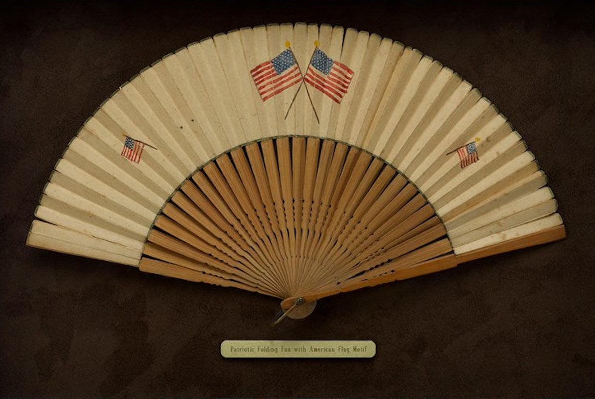 Presented is a patriotic folding paper fan, with an American flag motif. The fan is painted with two small American flags at left and right and two larger crossed American flags at center. The star count of the two crossed larger fans differ between