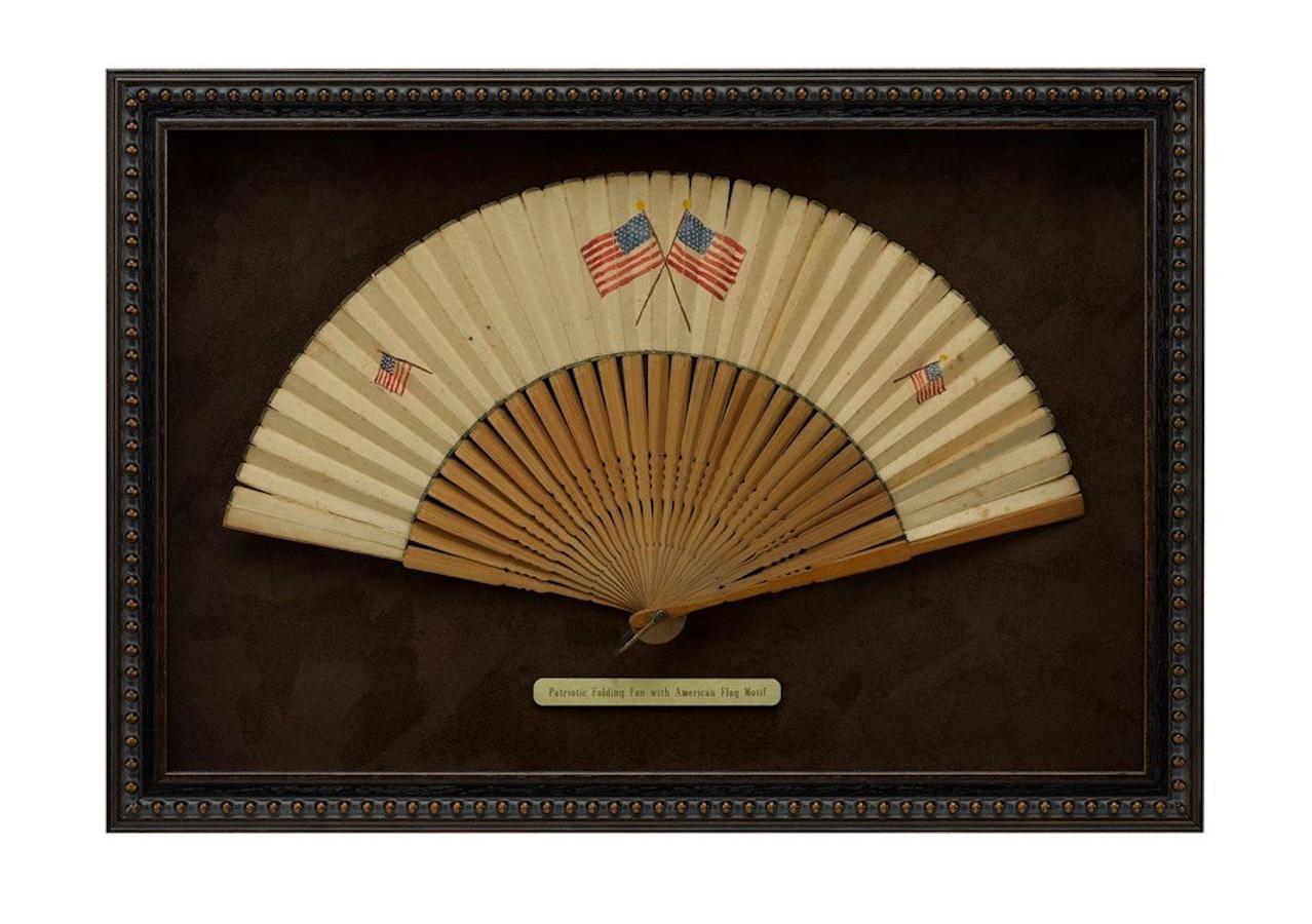Presented is a patriotic folding paper fan, with an American flag motif. The fan is painted with two small American flags at left and right and two larger crossed American flags at center. The star count of the two crossed larger fans differ between