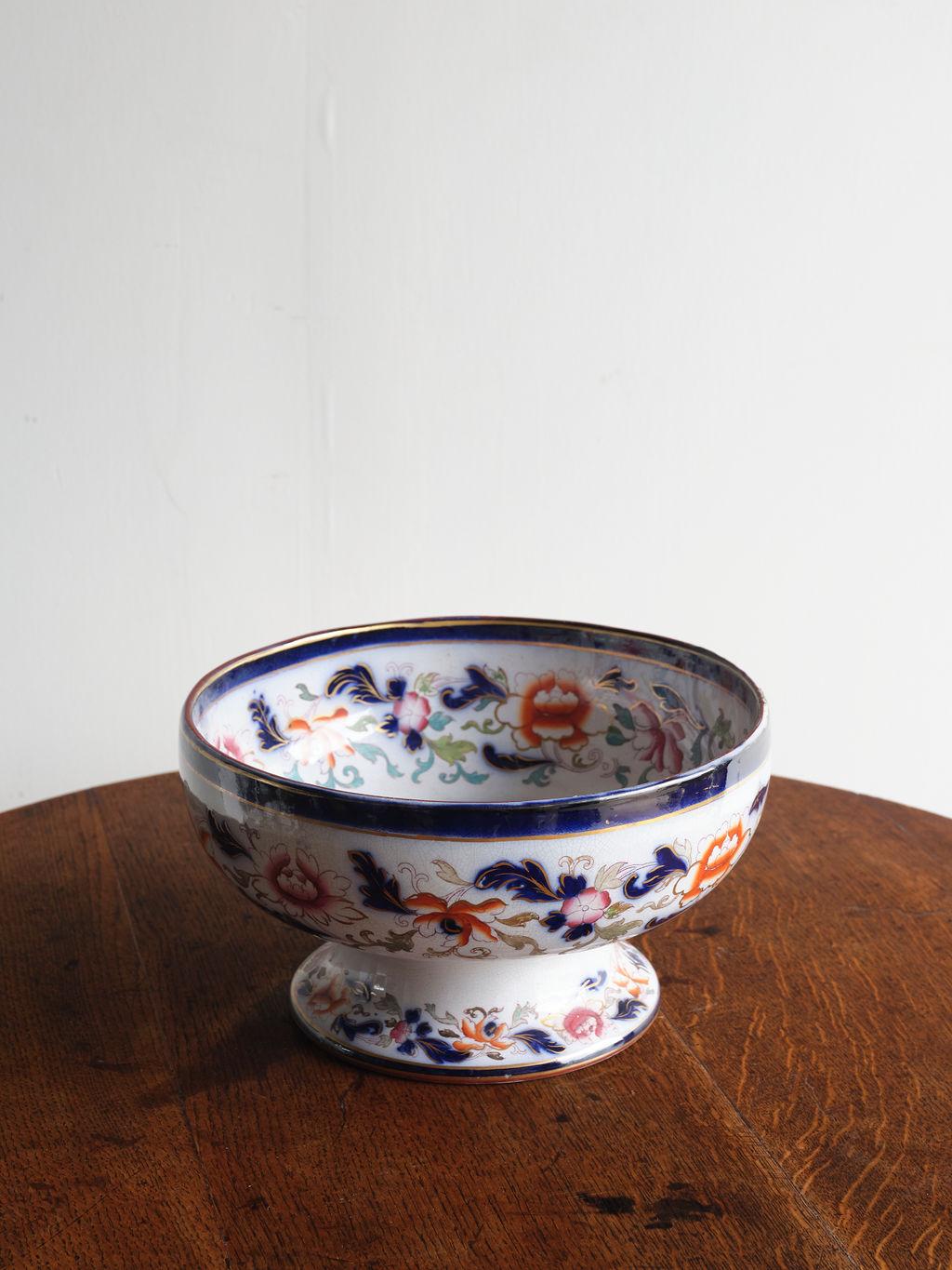 This lovely patterned ceramic bowl was crafted and hand painted in 1860 England. The floral details feature deep blue, dark orange and pink, and light tan and green coloring. There is a gold trim around the top and bottom of the dish. This bowl