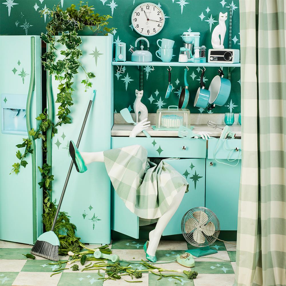 Atomic Kitchen de Patty Carroll présente une scène chaotique, une cuisine décorée de bleu sarcelle et d'aqua avec des plantes vertes et des produits qui débordent du réfrigérateur. Une femme semble être accaparée par cette cuisine, ses jambes