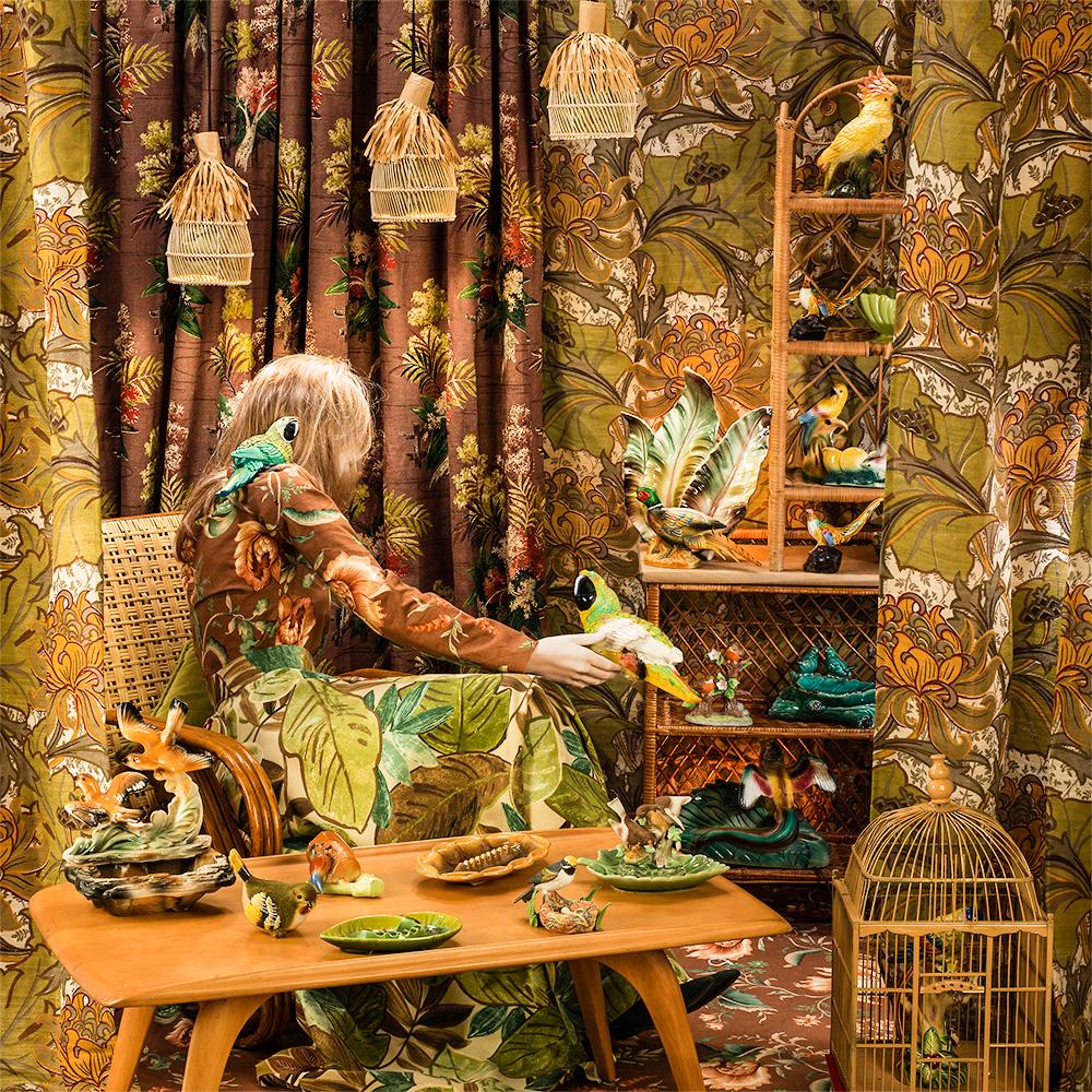 Bird in Hand by Patty Carroll presents a chaotic scene. A woman sits in a neutral toned room, surrounded by colorful birds. Botanical patterned curtains frame the figure. One bird rests on her shoulder while another sits on her hand. More birds sit