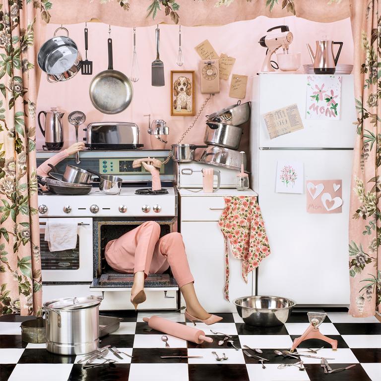 Cooking the Goose by Patty Carroll is a 38 x 38 inch archival pigment print. This photograph features a mannequin with arms and legs sticking out of an oven, surrounded by a mess of kitchen supplies. This print is signed, titled, dated and numbered