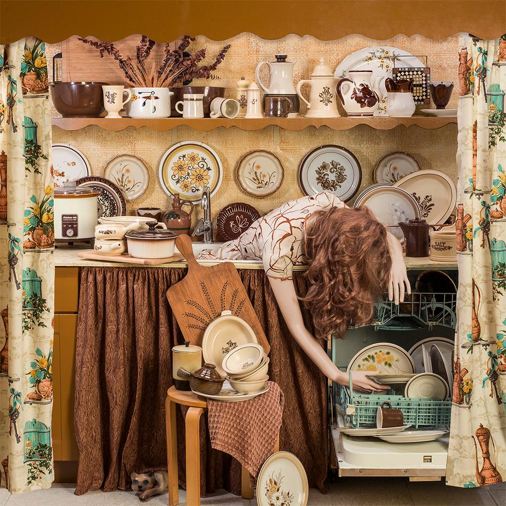 Duller Than Dishwater by Patty Carroll is an archival pigment print, available in an edition of 15. This photograph features a mannequin in the sink, reaching for dishes in the dishwasher. She is surrounded by neutral toned dishes, decor and drapes.